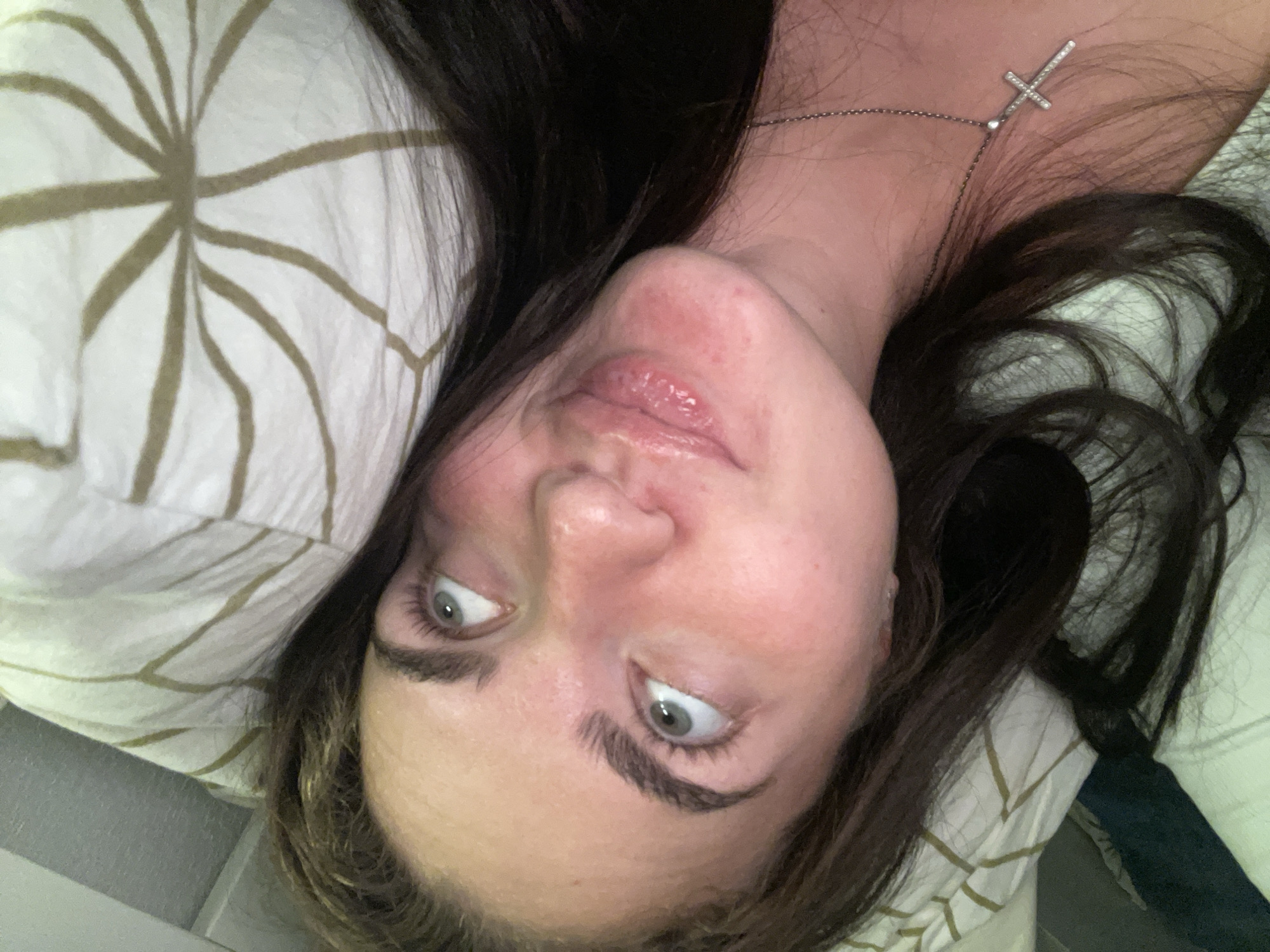 A young woman with dark hair, wearing a silver cross necklace, is lying in bed. She is looking up and to the right, with a slight smile. The background is a pillow with a brown and white geometric pattern. The scene appears to be indoors, likely in a bedroom. The location in Мосметаллоконструкция Онт, Russia, is not discernible from the image.