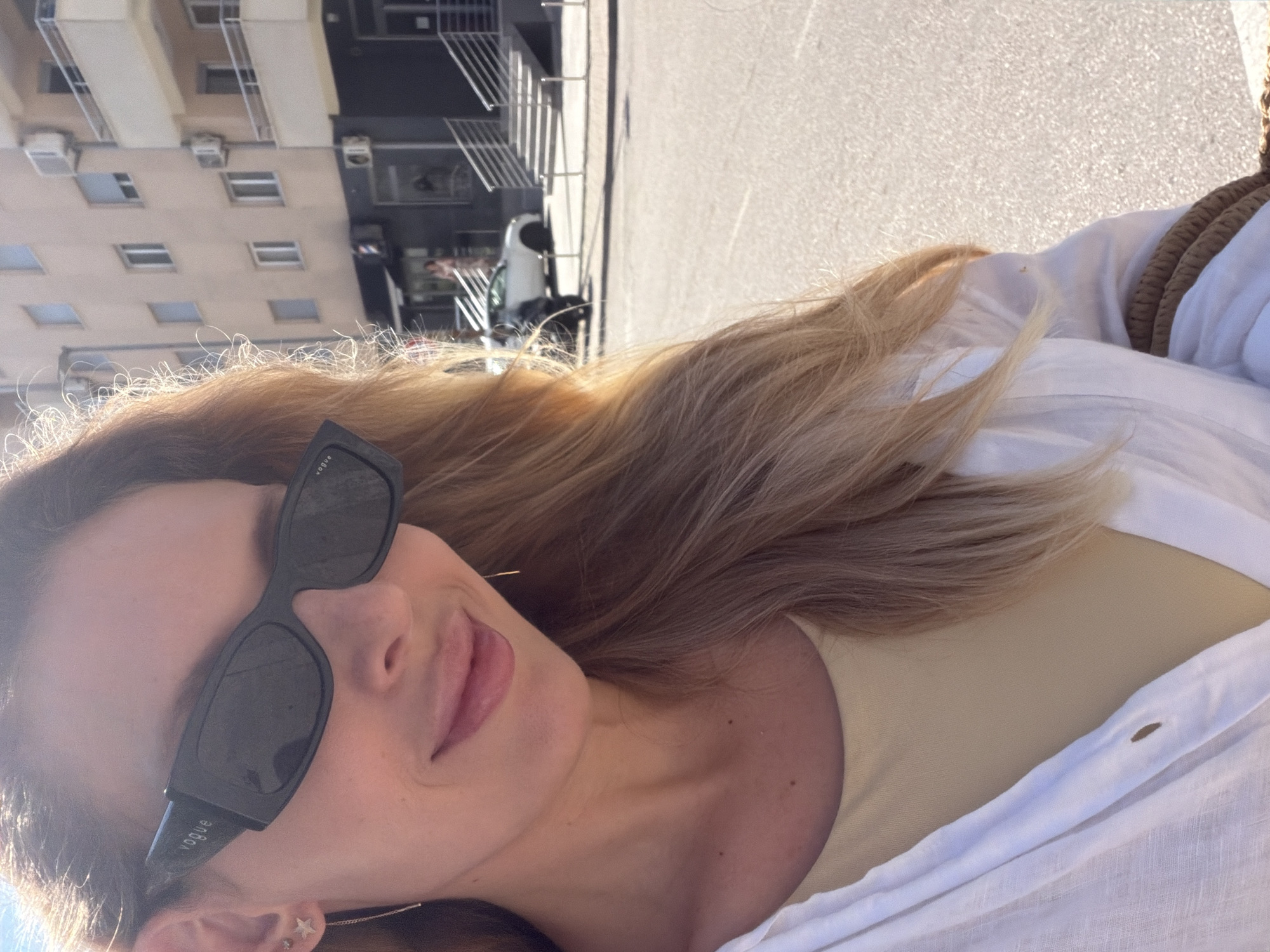 A woman with blonde hair, wearing black Vogue sunglasses, a light-colored top and a white open shirt, is standing outside on a sunny day in Budva, Montenegro. She is smiling and looking towards the camera. The background shows a building with balconies and windows, an air conditioning unit, a barber shop, a street with a white car parked, and a staircase leading up to an entrance. The ground is paved.
