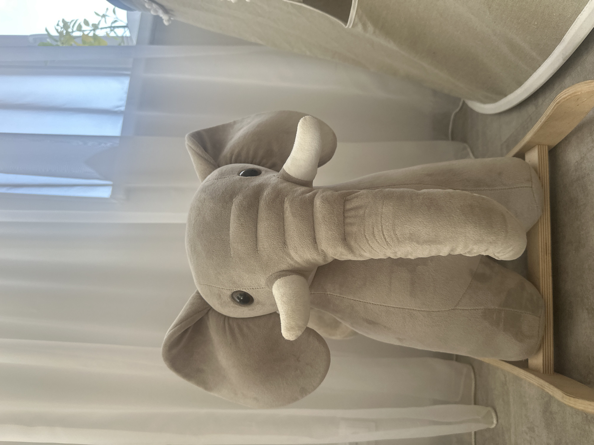 A close-up shows a stuffed, light brown plush elephant toy on a wooden rocking frame. The elephant has small white tusks and black button eyes. It appears to be located indoors. The backdrop consists of sheer white curtains and a portion of what looks like a fabric play tent or canopy. The specific location in the city of Гаревая, Russia, cannot be determined from the given visual information.
