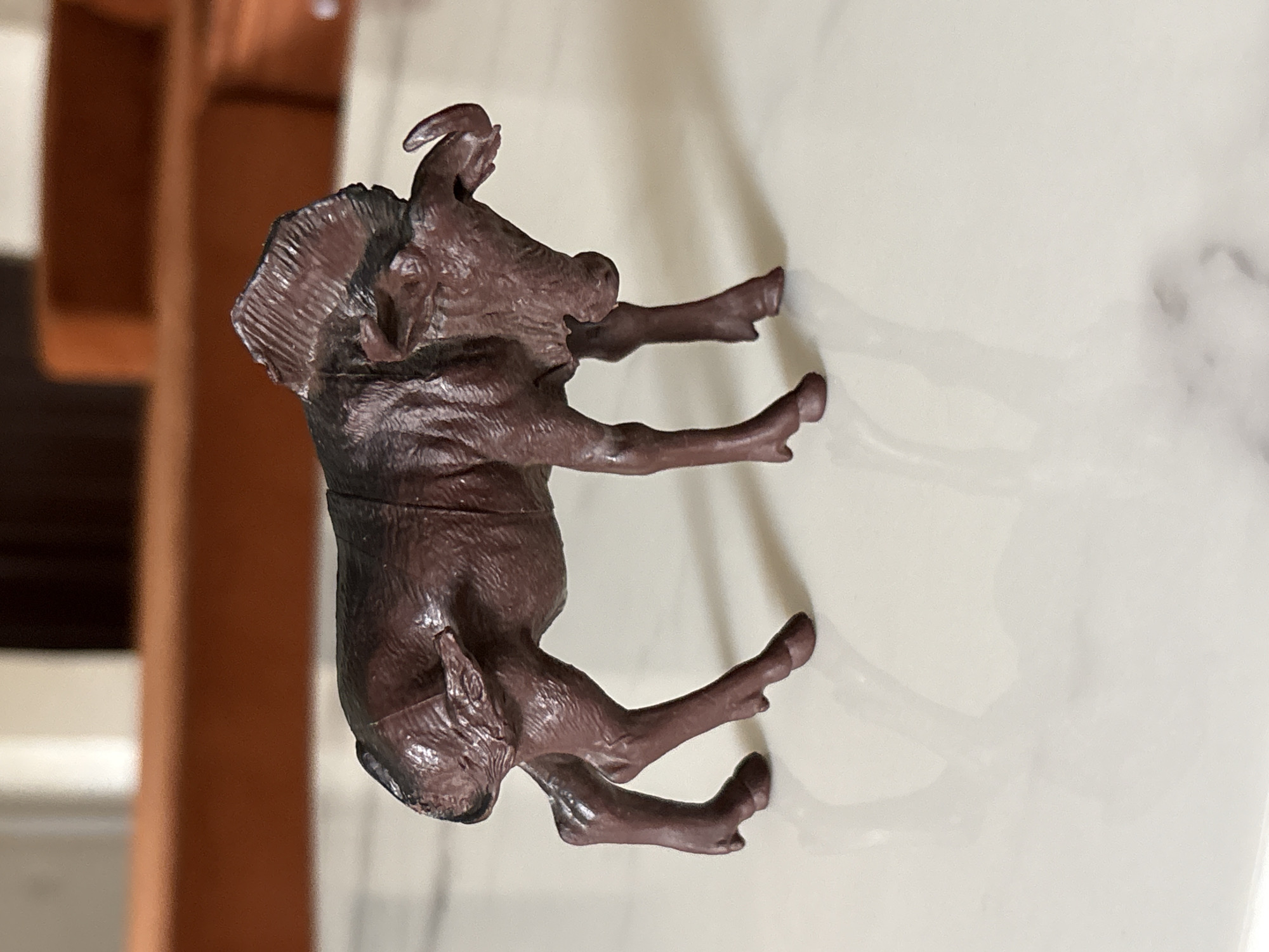 A dark brown, plastic or resin figurine, resembling a warthog or wild boar, is depicted horizontally across the frame. The figurine has a textured surface, mimicking wrinkled skin or coarse hair, with notable details including prominent ears, a snout, and two pairs of tusks or horns, one larger pair curving upwards from the top of its head, and a smaller set on its cheeks. Its four legs end in hoof-like feet. The figurine appears to be a toy or collectible.
It is positioned on a light, reflective surface, possibly marble or a glossy tile, which shows subtle variations in tone and some faint reflections. The background is softly blurred, displaying warm, rich brown hues, suggesting wooden furniture or a wall in an indoor setting. The lighting appears soft and even, highlighting the figurine's contours and textures. There are no people visible, nor any discernible activity taking place other than the static presence of the object. No text is visible in the image. The photo provides a close-up view of the figurine and its immediate surroundings.