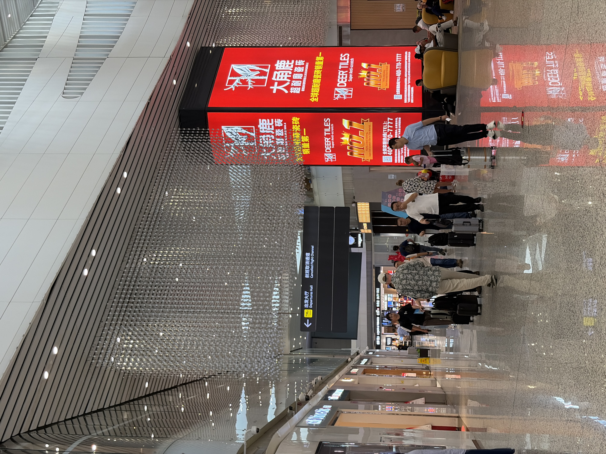 The image depicts the interior of Guangzhou Baiyun International Airport in Baiyun District, China. A large red advertisement for "Deer Tiles" is prominently displayed, showing both Chinese and English text and contact information. People are seen waiting with luggage, likely passengers. A sign indicates "Departures Hall" and "Cancelled Flight Channel." Shops are visible in the background. The ceiling has a complex, modern design.