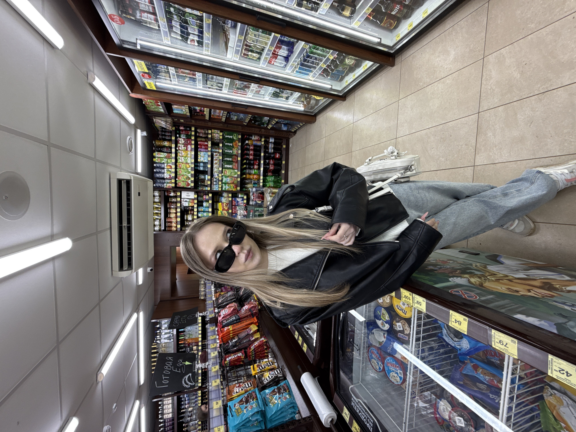The image, rotated 90 degrees counter-clockwise, shows a young woman posing casually in a grocery store aisle. She has long, straight blonde hair and is dressed in a black leather jacket over a white sweater, light blue jeans, and white sneakers. She wears black sunglasses and carries a small white handbag with a chain strap.
The background features typical supermarket elements: shelves densely stocked with various packaged goods, including candy brands like Skittles, M&M's, and Snickers, as well as drinks (some cans visible with "Rich" branding). Overhead, refrigerated display cases hold numerous beverages, while a large freezer chest in the foreground contains ice cream and other frozen products. Fluorescent lights illuminate the tiled floor and the store interior. An air conditioning unit is mounted on the wall.
Small blackboards with handwritten text in Russian are visible; one reads "Готовая Еда" (Ready Food) with a banana drawing, and another has "Есть" (Available). Other discernible Russian text includes "БЕЗ ЗАМЕНИТЕЛЯ" (without substitute) and "молочного жи" (dairy fat), likely from product labels. Price tags displaying numbers such as "42", "79", "84", "59", and "109" are affixed to shelves and freezers. A sign with "LIQUOR" is visible above a shelf. The scene captures a moment within a retail setting, characteristic of a grocery store in Russia.