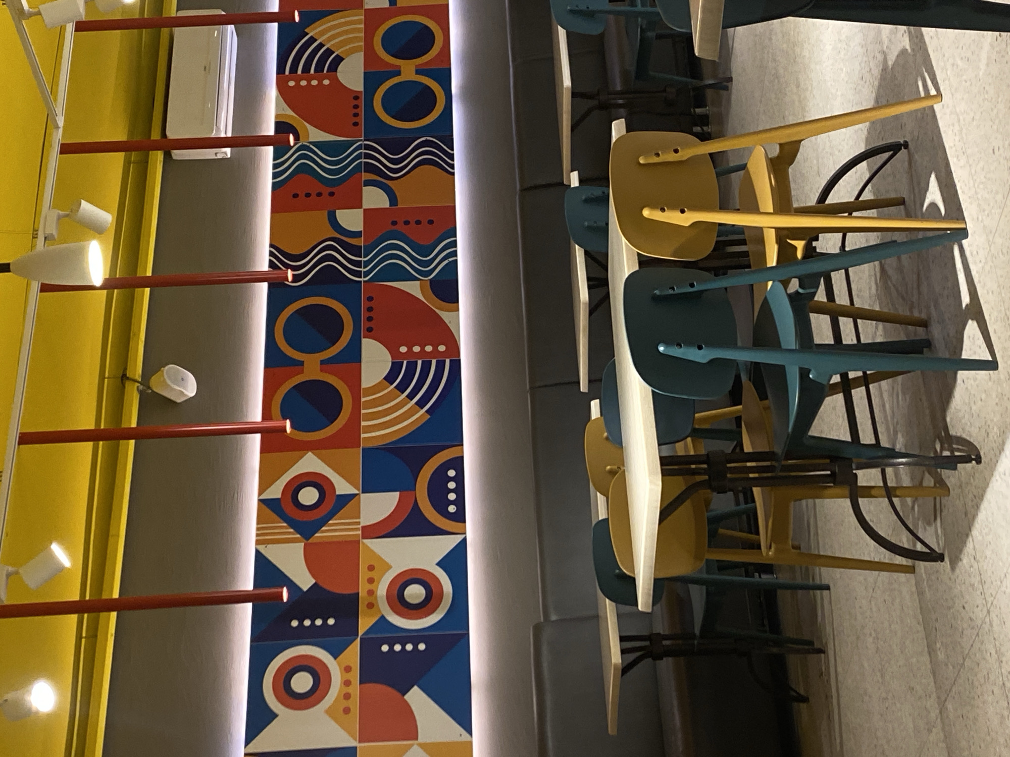The image depicts a section of a commercial interior, likely a restaurant or cafe, captured with the camera oriented sideways. The location is Berezniki, Russia.
On the left, a yellow wall contrasts with a grey column or wall section. A patterned, decorative tile design featuring geometric shapes and circles in red, orange, blue, and white is affixed to the wall. Track lighting with red rods and spotlights illuminates the scene. An air conditioning unit is mounted high on the gray wall.
On the right, stacked chairs in yellow and blue-green are visible. The chairs appear to be stacked on a narrow ledge or platform. The floor beneath the chairs is lightly textured. A padded bench, in grey, is running in the background.