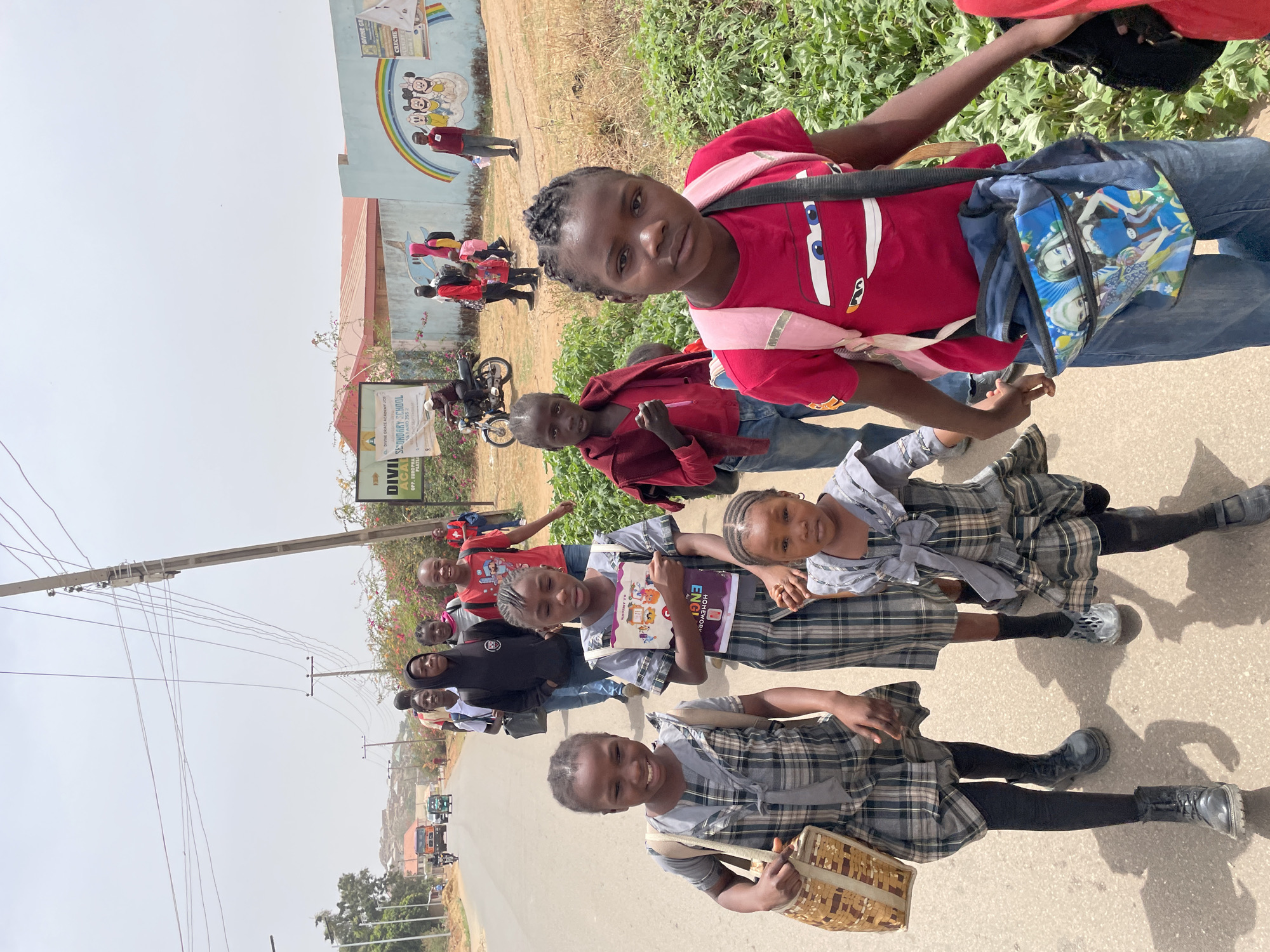 A medium-shot, slightly elevated outdoor photograph captures a group of children, predominantly school-aged, walking along a paved road under bright daylight in Jos, Nigeria. The clear, hazy sky and visible shadows suggest a sunny day, likely mid-morning or afternoon.
In the foreground, several girls are prominent. Two girls wear plaid school uniforms (skirts/dresses) with light-colored tops, black tights, and shoes or boots. One of them holds a light-colored woven basket, while another, slightly behind, holds a book titled "HOMEWORK ENGLISH". Further back, a girl in a red t-shirt, featuring a "Cars" character (Lightning McQueen), wears a pink sash and a blue backpack adorned with "Frozen" characters. Other children and a few adults are visible further down the road, some also in school uniforms, others in casual wear like red shirts and jeans. Most of the children are looking towards the camera, with some smiling and others having neutral or serious expressions.
The background features a building with a colorful mural depicting Disney characters, including Mickey Mouse, Minnie Mouse, Pluto, and Goofy. Near this building, a green sign, partially visible, reads "SECONDARY SCHOOL" and includes "DIVINE ACADEMY" and "CRICHOL," along with an address like "OPP. EURO PEAS" and "PLOT B164." A motorcycle is parked near the sign, and power lines stretch across the upper left portion of the frame. The scene portrays children likely on their way to or from school.