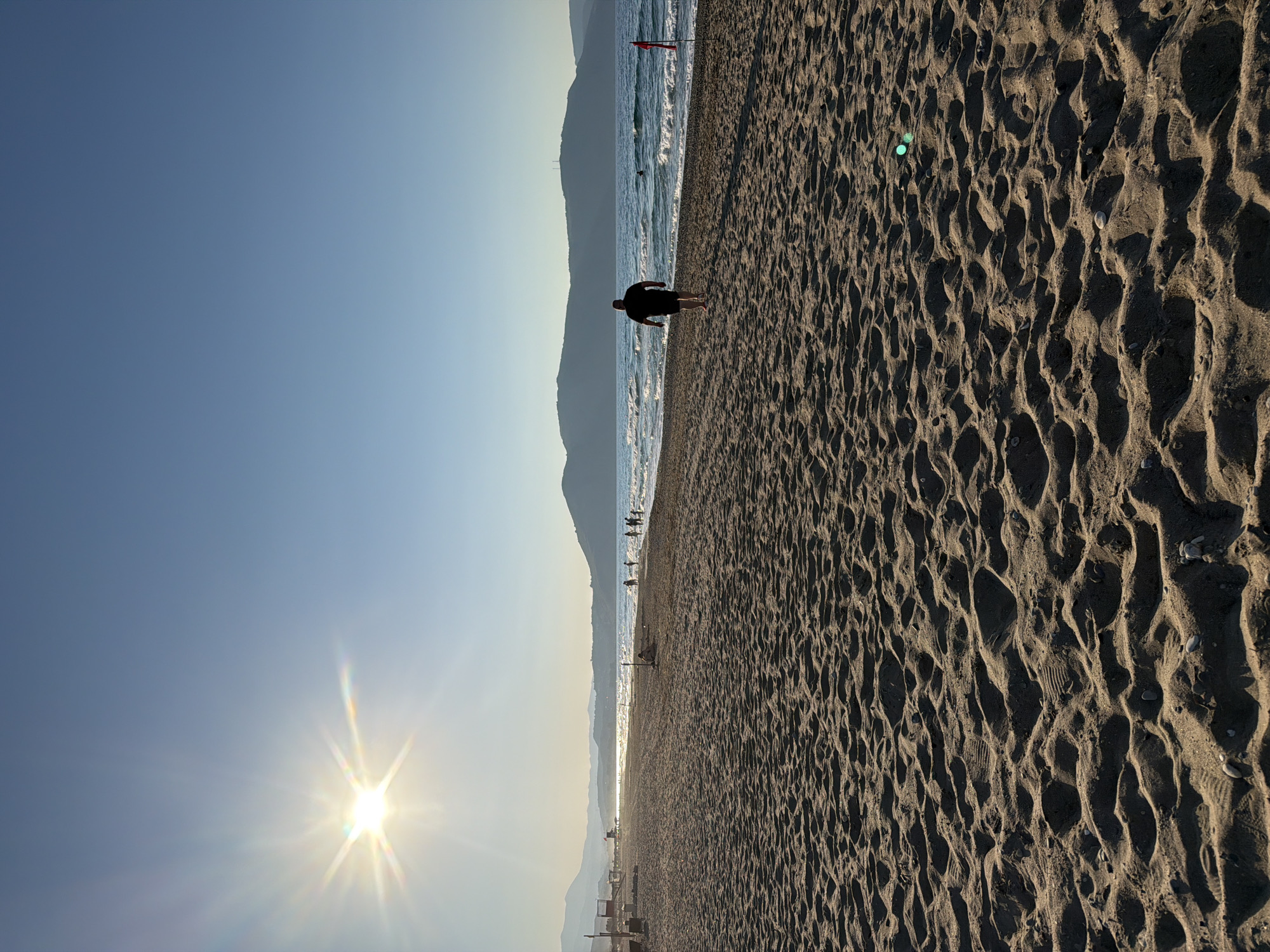 The image depicts a wide, sandy beach in Dramia, Greece, under a clear blue sky. The sun is shining brightly, casting long shadows across the rippled sand in the foreground. In the middle ground, several people are walking along the shoreline or wading in the gentle waves of the sea. The water appears calm with small whitecaps. In the background, rolling hills or mountains form a distant horizon. A single person, a male wearing dark shorts and a dark t-shirt, is walking away from the camera towards the sea. There are also a few beach structures visible in the far distance. The overall scene is tranquil and evokes a sense of a late afternoon at the beach.