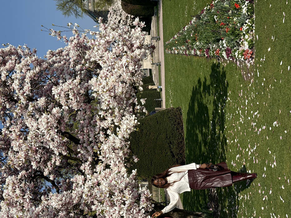 The image depicts an outdoor scene in Parc de l'Orangerie, Strasbourg, France, on a clear, sunny day. A woman is positioned in the left foreground, leaning her left arm against the trunk of a large magnolia tree in full bloom with numerous pink and white flowers. She has dark skin and long dark hair, wears a white long-sleeved blouse, a dark reddish-brown midi-length skirt, and matching knee-high boots, holding a dark handbag in her right hand. Her head is turned slightly to her right, and she exhibits a subtle smile.
The ground consists of neatly manicured green grass, with numerous fallen magnolia petals scattered across it, particularly in the foreground and midground. A linear flower bed containing various red, white, and purple flowers runs diagonally from the mid-ground right towards the foreground.
In the background, further green hedges and trees are visible. Two distant figures are present; one person in a light-colored coat is standing on a paved area, facing away from the viewer, beyond the flower bed. Another person is partially obscured by a hedge on the left background. A large, light-colored building with classical architectural elements, including columns, is partially visible in the far background on the right. The sky above is clear and blue. Shadows are cast by the trees, indicating bright sunlight.