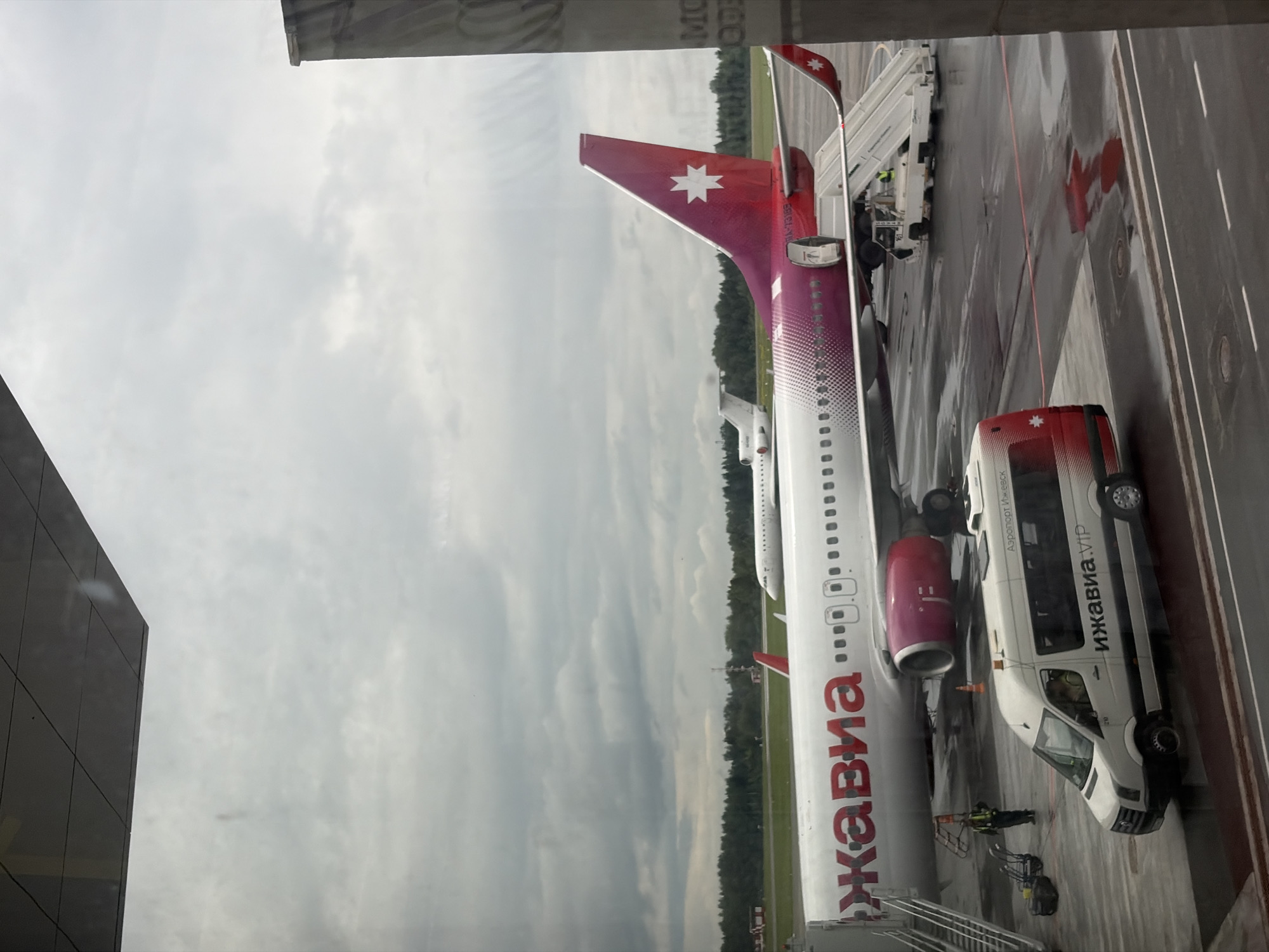 The image captures an airport scene, likely viewed from within a terminal building through a window, looking out onto the apron. The sky is overcast and cloudy, suggesting a daytime setting with no direct sunlight. The tarmac below appears wet in several areas, possibly from recent rain or cleaning.
The primary subject is a passenger aircraft, identifiable as belonging to the "ижавиа" (Izhavia) airline, based on the red Cyrillic text on its white fuselage. The plane features a distinctive red and magenta tail fin with a white star logo, and its engines are also magenta. The aircraft registration "RR-73062" is visible on the tail.
Adjacent to the plane, a white airport shuttle bus is parked, also bearing "ижавиа.VIP" and "Аэропорт Ижевск" (Izhevsk Airport) text on its side, confirming the location and airline. "Sprinter" is also visible on the front of the bus. Mobile passenger stairs are connected to the aircraft, indicating ongoing ground operations such as boarding or disembarkation. At least two airport ground crew members are visible near the aircraft and equipment, engaged in their duties.
In the background, a distant treeline separates the airport area from what appears to be a natural or less-developed landscape. The visible texts "Izhavia" and "Izhevsk Airport" strongly suggest the location is Izhevsk Airport, situated near Новая Казмаска, Russia.