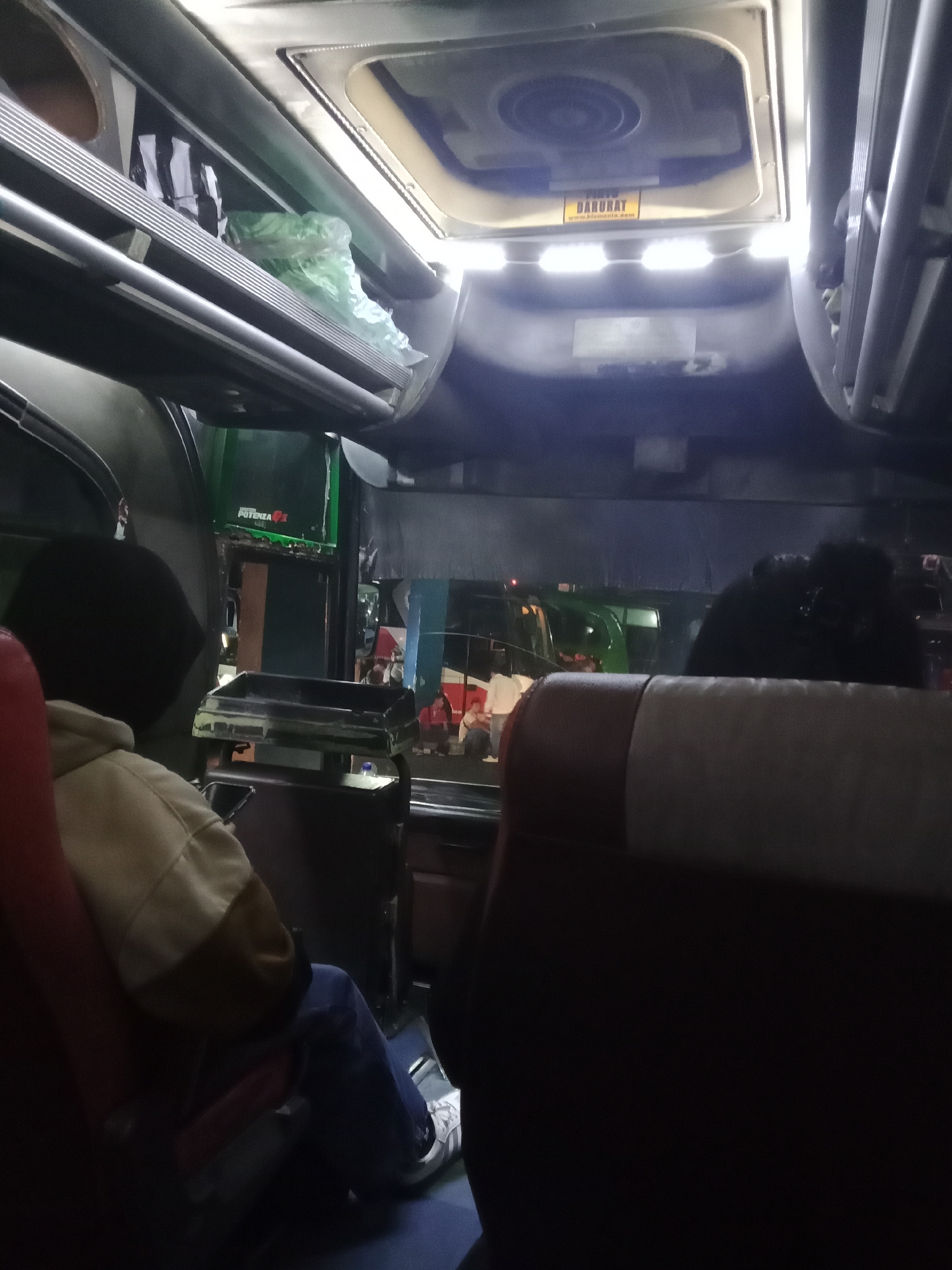 The image is an interior view of a bus, likely in Surakarta, Indonesia. The perspective is from a passenger seat, looking forward and slightly to the left. In the foreground, a person wearing a dark hooded jacket and blue jeans is seated, with their left leg visible. The bus cabin features overhead luggage racks with some items stored on them, including a green plastic bag. A ventilation or air conditioning unit is visible on the ceiling, with a yellow sticker that reads "DARURAT" and a website address. Further ahead, other passengers are visible in their seats, some in profile or from behind. Outside the bus windows, other vehicles and some structures are faintly discernible, suggesting a street scene. The lighting is dim, characteristic of an indoor environment at night or during low light conditions.