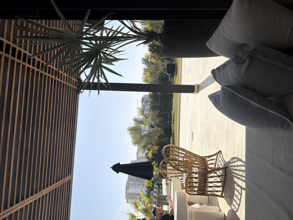 The image depicts an outdoor lounge area or patio, likely associated with a restaurant, cafe, or hotel, situated in an urban part of Dubai, United Arab Emirates. The scene is illuminated by bright sunlight under a clear blue sky.
In the immediate foreground on the right, light-colored lounge cushions and several pillows (one white, two grey/blue) are visible, indicating a seating arrangement. The perspective is from this low seating position.
To the left of the frame, a male individual wearing a black t-shirt is seated at a round white pedestal table, looking downwards, with his right hand near his face. Opposite him, a woven rattan chair with a curved back stands, casting a pronounced shadow on the light-colored paved patio. Another similar white table is partially visible to the left.
A closed black patio umbrella is positioned centrally in the mid-ground. Beyond the patio, numerous fan palm trees, planted in large dark containers, are distributed across a landscaped area with green grass, separated by a low fence. In the distant background, multiple light-colored, multi-story buildings with balconies are visible, characteristic of residential or commercial structures in Dubai. A construction crane is observable above the buildings on the right horizon.
Overhead, a wooden slatted pergola provides partial shade to the patio area. The strong sunlight casts distinct shadows across the patio.