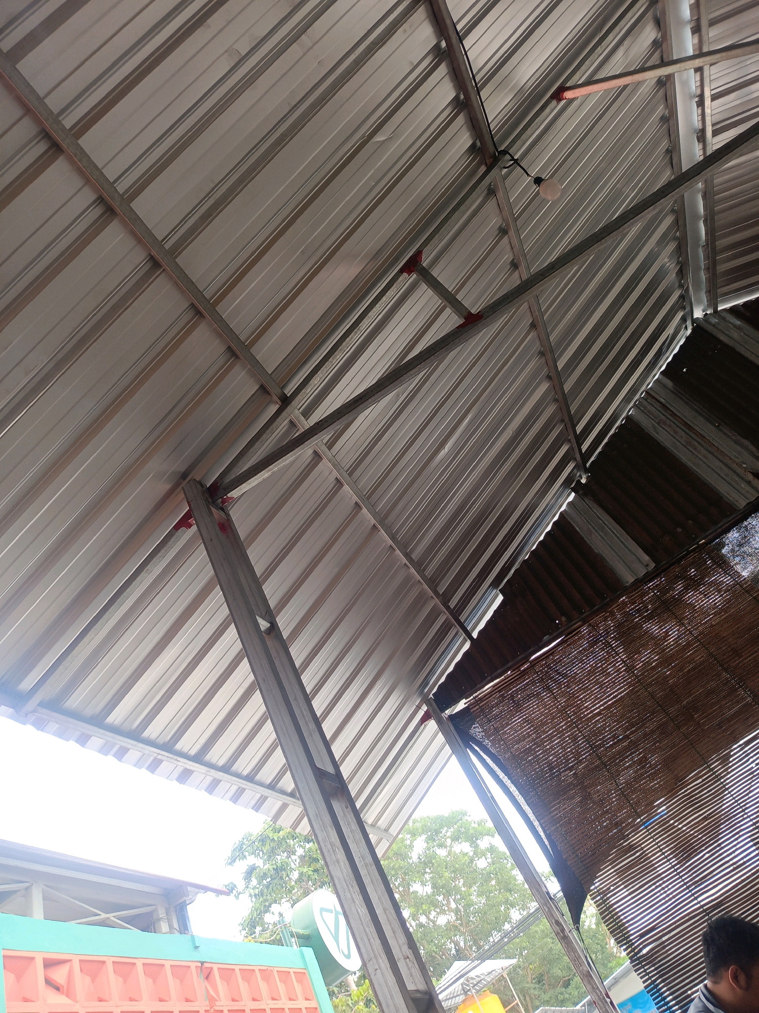 The image shows the underside of a metal roof supported by metal beams. The roof is corrugated and light gray. A simple incandescent light bulb hangs from a wire. There are also red brackets visible holding some of the beams. To the right, a woven bamboo blind partially covers the view, and further to the right, a portion of a person's head and shoulder is visible. In the background, through gaps in the roof and blind, trees and a bright sky are visible, suggesting an outdoor or semi-outdoor setting. The scene appears to be a covered structure, possibly a market stall or open-air dining area, in Magetan, Indonesia.