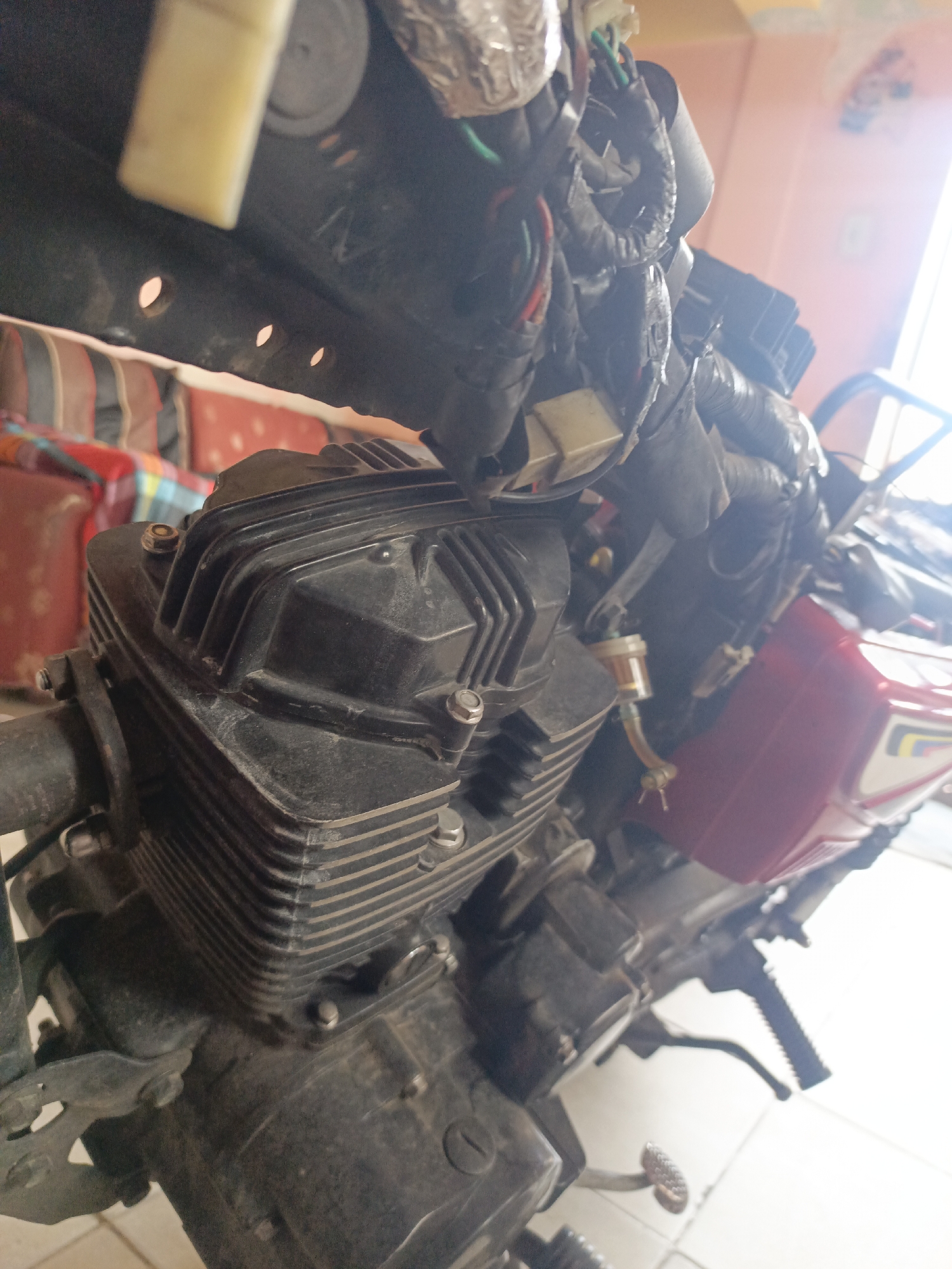 The media file shows a close-up view of a motorcycle engine and wiring. The engine is black with prominent cooling fins, and a small fuel filter is visible attached to a hose. A red motorcycle body panel with some white and yellow decals is also visible to the right of the engine. The wiring harness, wrapped in black tape, is clustered at the top of the engine. The scene appears to be indoors, possibly a workshop or garage, with a bright light source coming from the right, suggesting daytime. The background shows parts of what appears to be furniture with colorful striped upholstery, and a light-colored wall. There are no people visible in the image. The overall impression is that of a partially disassembled or maintained motorcycle. There is no visible text in the image. The location context is Juan Griego, Venezuela.