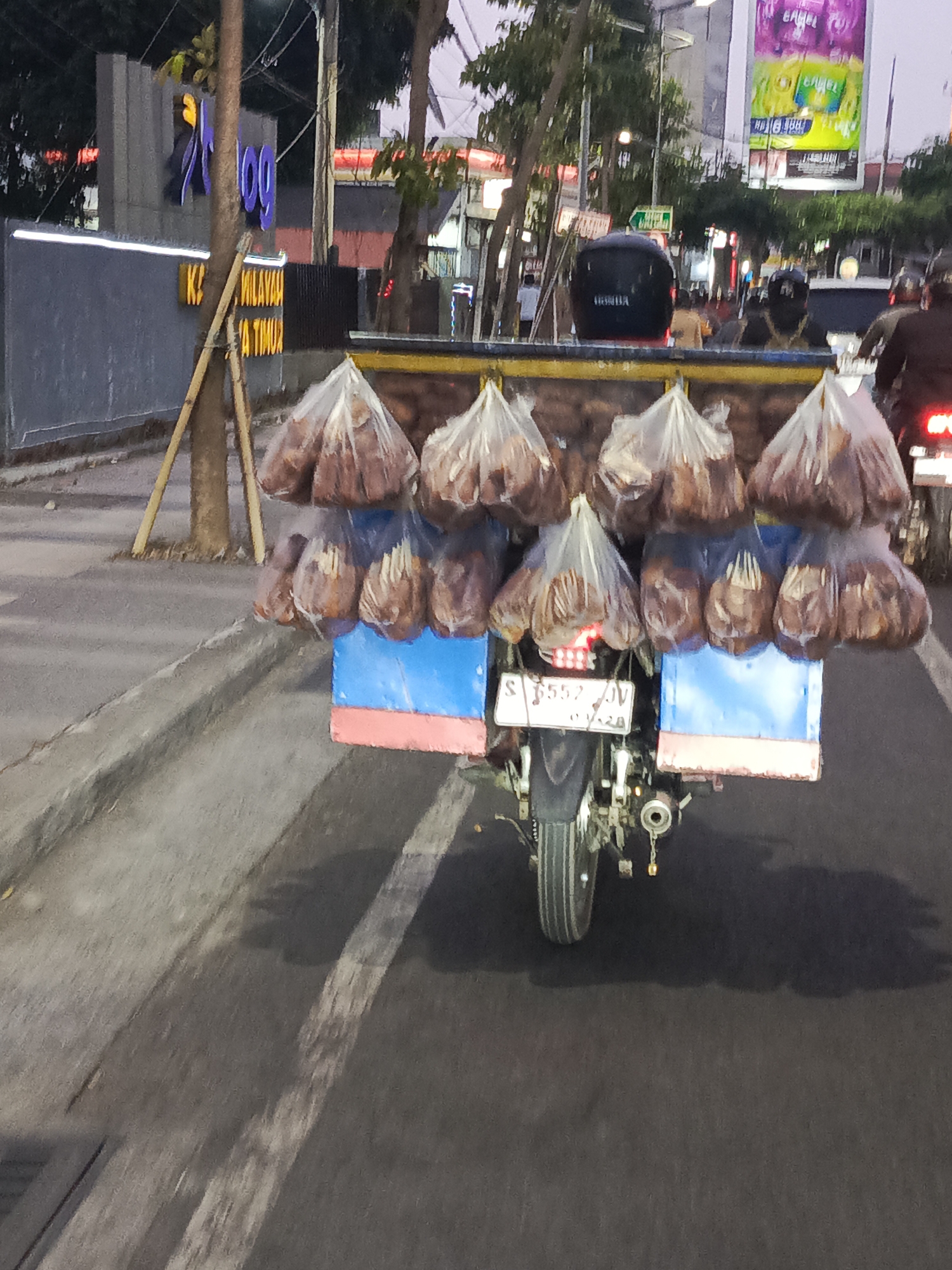 The image shows a street scene in Surabaya, Indonesia, likely in the evening. A motorcycle is seen from behind, heavily laden with goods in plastic bags. The goods appear to be food items, possibly snacks or produce, packed in numerous transparent plastic bags hanging from a metal rack attached to the back of the motorcycle. The motorcycle has a license plate that reads "S 5557 DV".
The background features urban elements: buildings with signage, streetlights, and trees. One prominent sign in the distance appears to be an advertisement with colorful graphics. The street is paved and has a curb. Shadows are cast on the road, suggesting the sun has set or is low in the sky. A few other people are visible in the distance, some on motorcycles, indicating traffic on the street. The overall atmosphere is that of a typical street in a bustling Indonesian city.