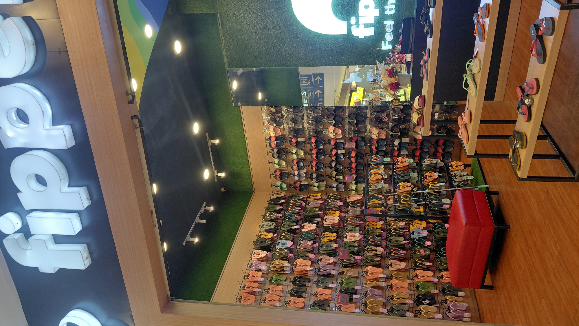 The image shows the interior of a Flipper shoe store, likely located in the city of Purwokerto, Indonesia. The store features walls lined with rows of flip-flops in various colors and styles, displayed on metal racks. In the foreground, there are several display shelves with more flip-flops arranged on them. A red ottoman or seating bench is positioned to the left of the frame. The store has a modern design with wooden accents and a black ceiling with track lighting. The Flipper logo is visible above the entrance. A mirror on the right wall reflects a portion of the store and the mall environment beyond. There are no people clearly visible in the image, suggesting it was taken during a quiet period or when the store was not busy. The overall impression is of a well-organized retail space showcasing footwear.