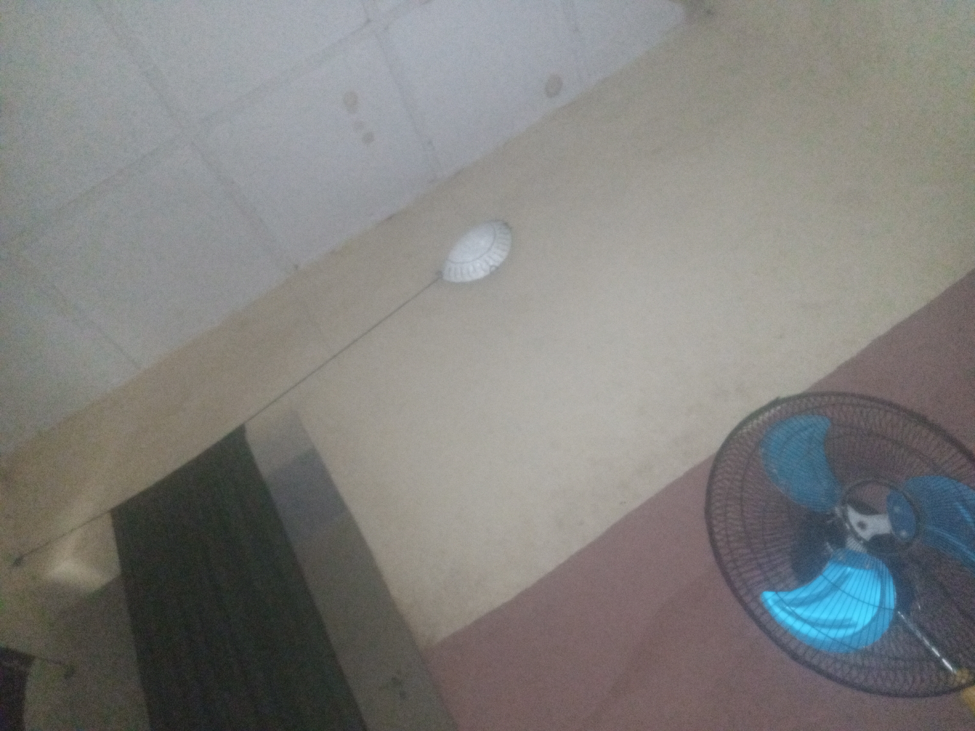 The image depicts an indoor scene. In the upper portion of the frame, a white, circular object is suspended from the ceiling by a thin wire. This object appears to be a smoke detector or a similar ceiling-mounted device. Below and to the right of this device, a large black electric fan with bright blue blades is prominently displayed. The fan is positioned on a surface that appears to be a table or a floor. The walls are a light beige color, and the ceiling is white with visible seams. The overall lighting is dim, suggesting it might be nighttime or the room is not well-lit. There are no people or animals visible in the image.