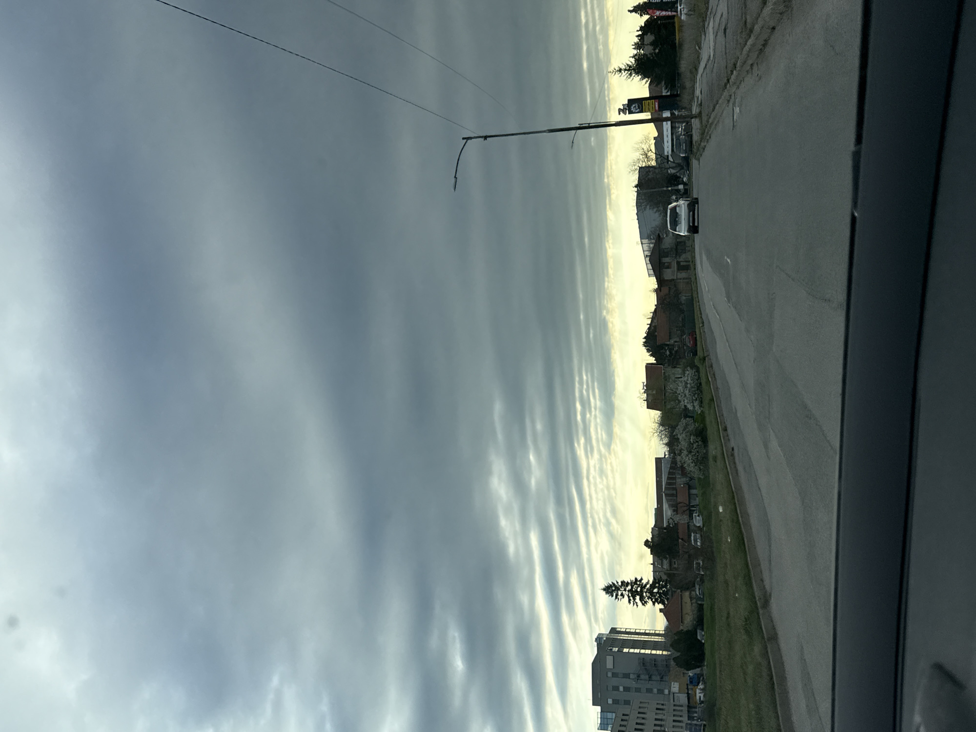 Captured from inside a vehicle traveling on a gray asphalt road, the image presents a cityscape under an overcast sky patterned with distinct, parallel cloud formations. A single white vehicle is visible ahead on the road.
On the right side of the road, a grassy verge precedes a row of buildings of varying heights and designs, including a tall, modern building with multiple stories. Mature trees are scattered among the buildings. Streetlights are spaced along the road.
The scene appears to be in Sofia, Bulgaria, based on the architecture and overall urban layout. The image suggests an early morning or late afternoon due to the soft light. The sky is dominated by a rippled cloud pattern, possibly altocumulus undulatus.