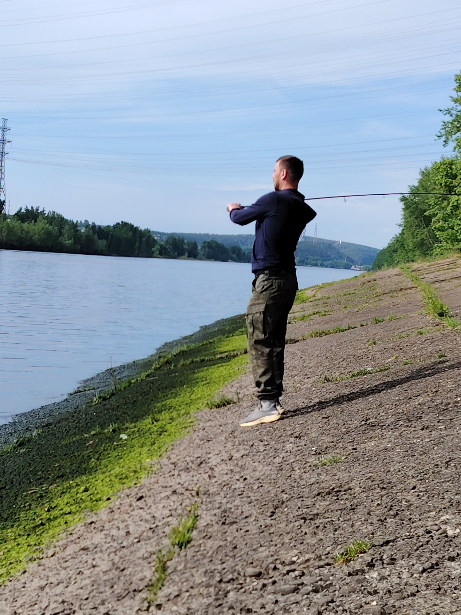 A man stands fishing on a concrete embankment along a river in Chaikovsky, Russia. He is facing away from the viewer, wearing a dark blue hooded sweatshirt, camouflage cargo pants, and gray sneakers with tan soles. He holds a fishing rod with both hands. The river is to his left, lined with green vegetation along the edge. In the distance across the river, forested hills are visible. Power lines and a tower are present in the sky. The concrete embankment is cracked and covered with patches of green moss and sparse grass.