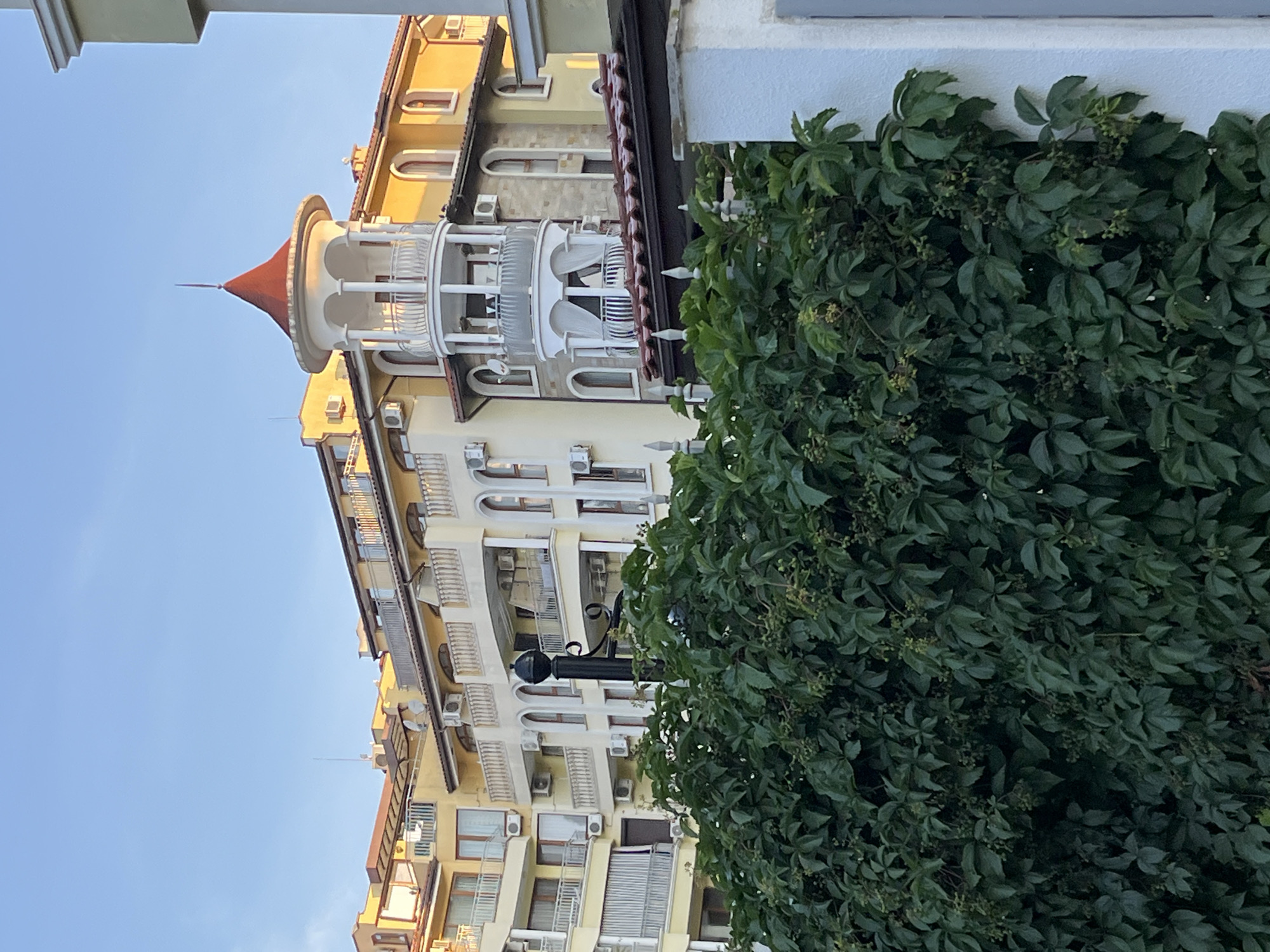 The image shows a multi-story building in Nesebar, Bulgaria, characterized by its yellow facade and white balconies. A prominent feature is a circular tower with a red conical roof, adorned with white columns and railings. The building is partially obscured by lush green foliage in the foreground, which appears to be a dense vine covering a wall or fence. The sky above is a clear, pale blue, suggesting it is daytime. The architecture suggests a hotel or residential complex.