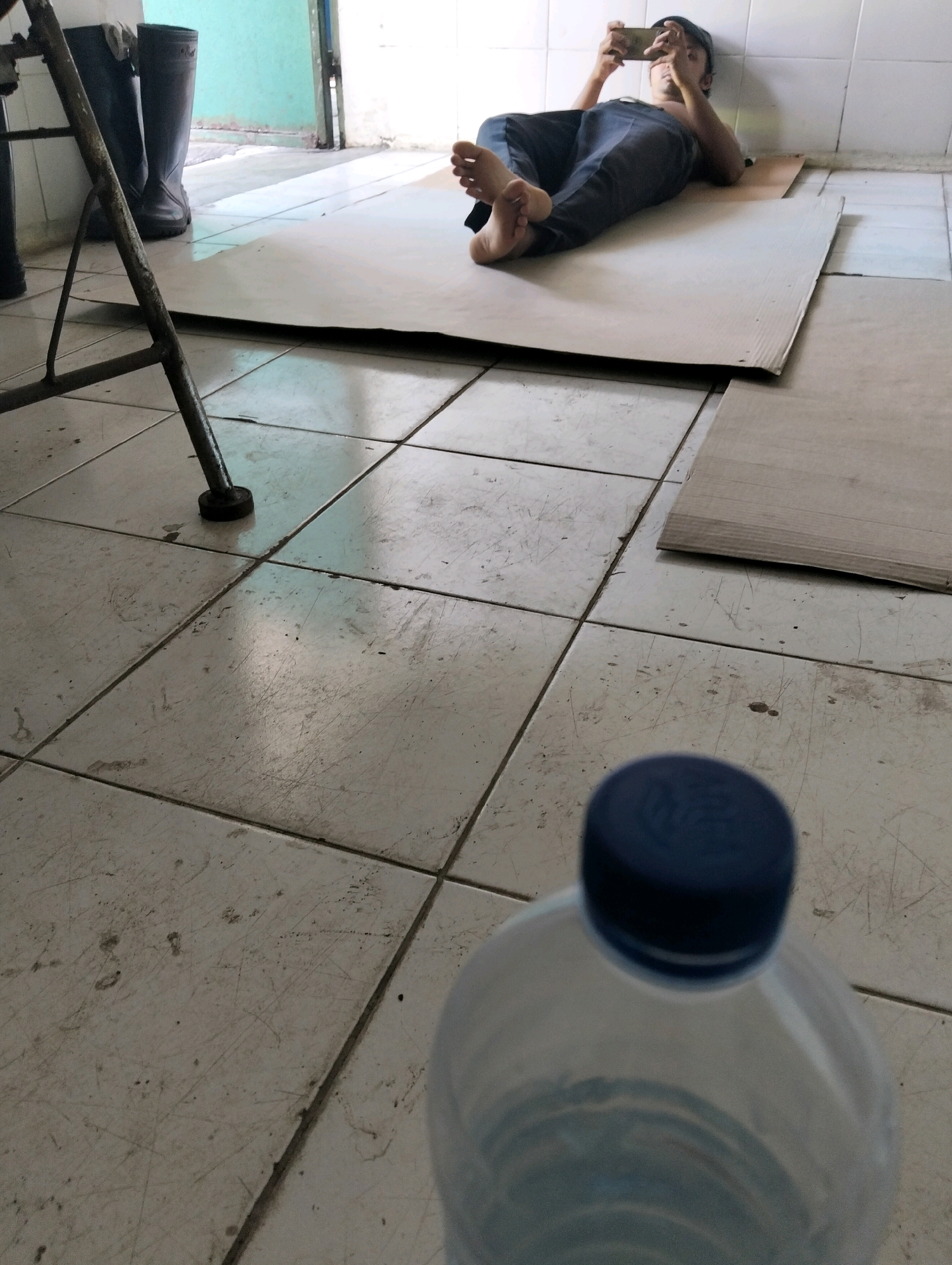 A person lies on cardboard placed on a tiled floor, using a mobile phone. They are wearing dark pants and a dark cap. Their feet are bare. Two dark boots are visible to the left of the frame, and a portion of a metallic stand is also present. In the foreground, the blurred top of a plastic water bottle with a blue cap is visible. The environment appears to be indoors, possibly a utilitarian space with light-colored walls and a tiled floor. The scene is set in Tanggulangin, Indonesia.