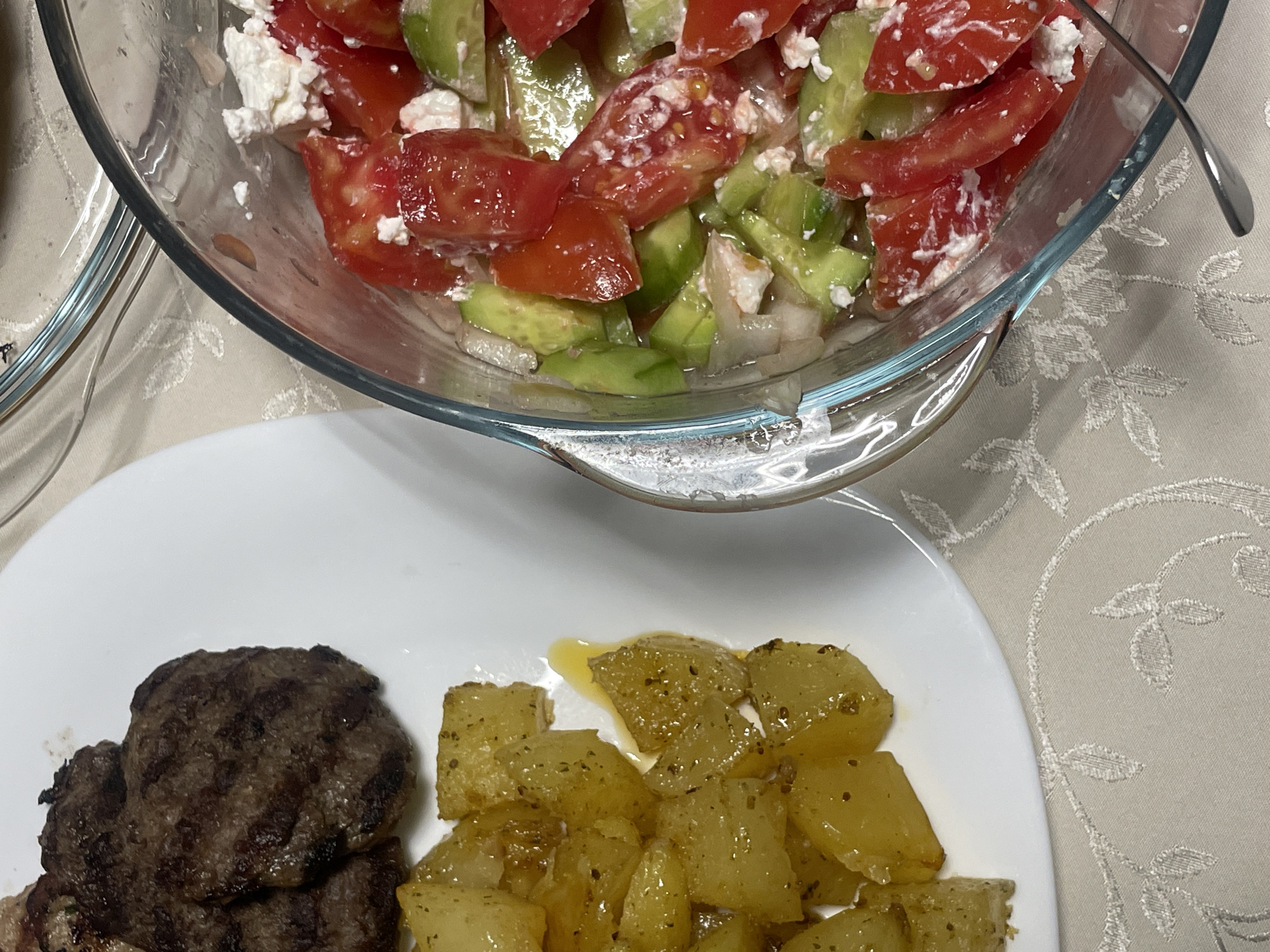 A close-up shot shows a meal consisting of grilled meat patties, seasoned potatoes, and a tomato and cucumber salad with feta cheese. The food is arranged on a white plate and in a glass bowl, placed on a textured tablecloth. The scene appears to be indoors, possibly in a dining area. No specific location in Velingrad, Bulgaria, is discernible from the image itself.
