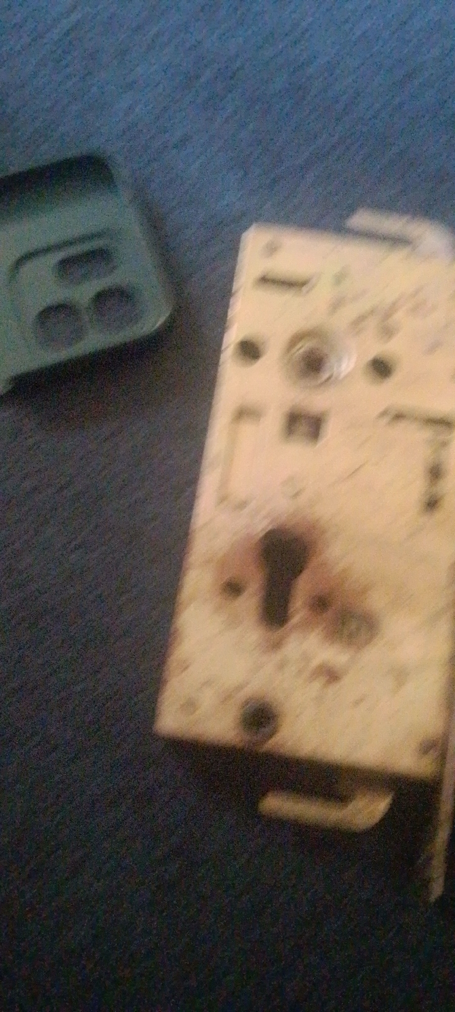 The image shows a close-up, out-of-focus view of a lock mechanism on a dark, textured surface, likely a carpet or fabric. To the left, a green object, possibly a phone case, is partially visible. The lock mechanism is metallic, with visible screws, a keyhole, and some discoloration or wear. The lighting appears to be dim, contributing to the overall blurriness of the image. There are no people or discernible actions taking place. The scene is devoid of any specific geographical or urban indicators, making the location "Unknown, Unknown."