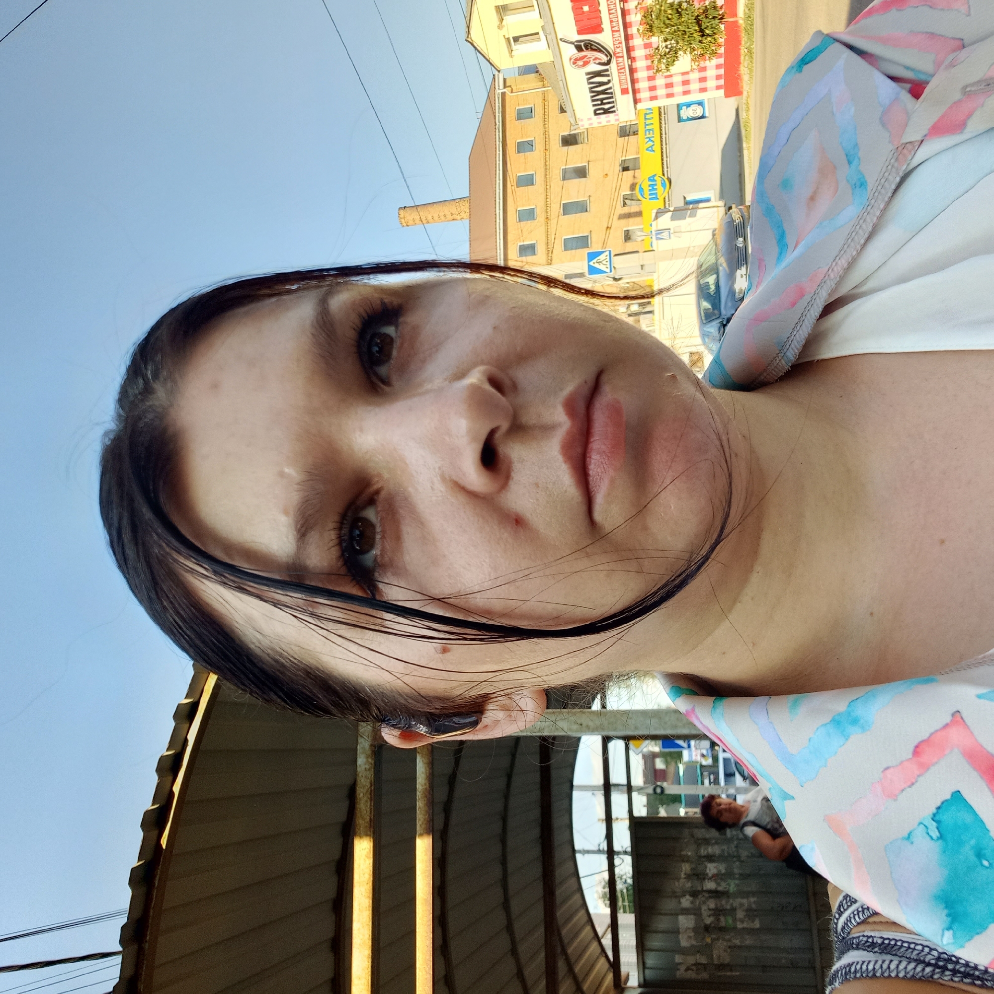 A close-up, waist-up, outdoor, daytime selfie shows a woman with dark hair pulled back and some strands falling forward. She is wearing a white top and a patterned jacket with watercolor-like designs in pink, blue, and teal. She has a black earbud in her left ear. Her expression is neutral, and she is looking slightly upward and to her right.
The background suggests an urban setting in Kropyvnytskyi, Ukraine, likely near a bus stop or public shelter. To the left, a corrugated metal structure with horizontal beams and a translucent roof is visible. Behind her, a street is visible with cars, buildings, and signage. One building is a light brown, multi-story structure with numerous windows, and a tall, narrow brick chimney stands next to it. Another building to the right is yellow and has white window frames. A red and white checkered awning is visible on a building facade. A sign with Cyrillic lettering is present. The sky is clear and blue. In the middle ground, a woman in a white shirt is standing, partially obscured by the woman in the foreground.