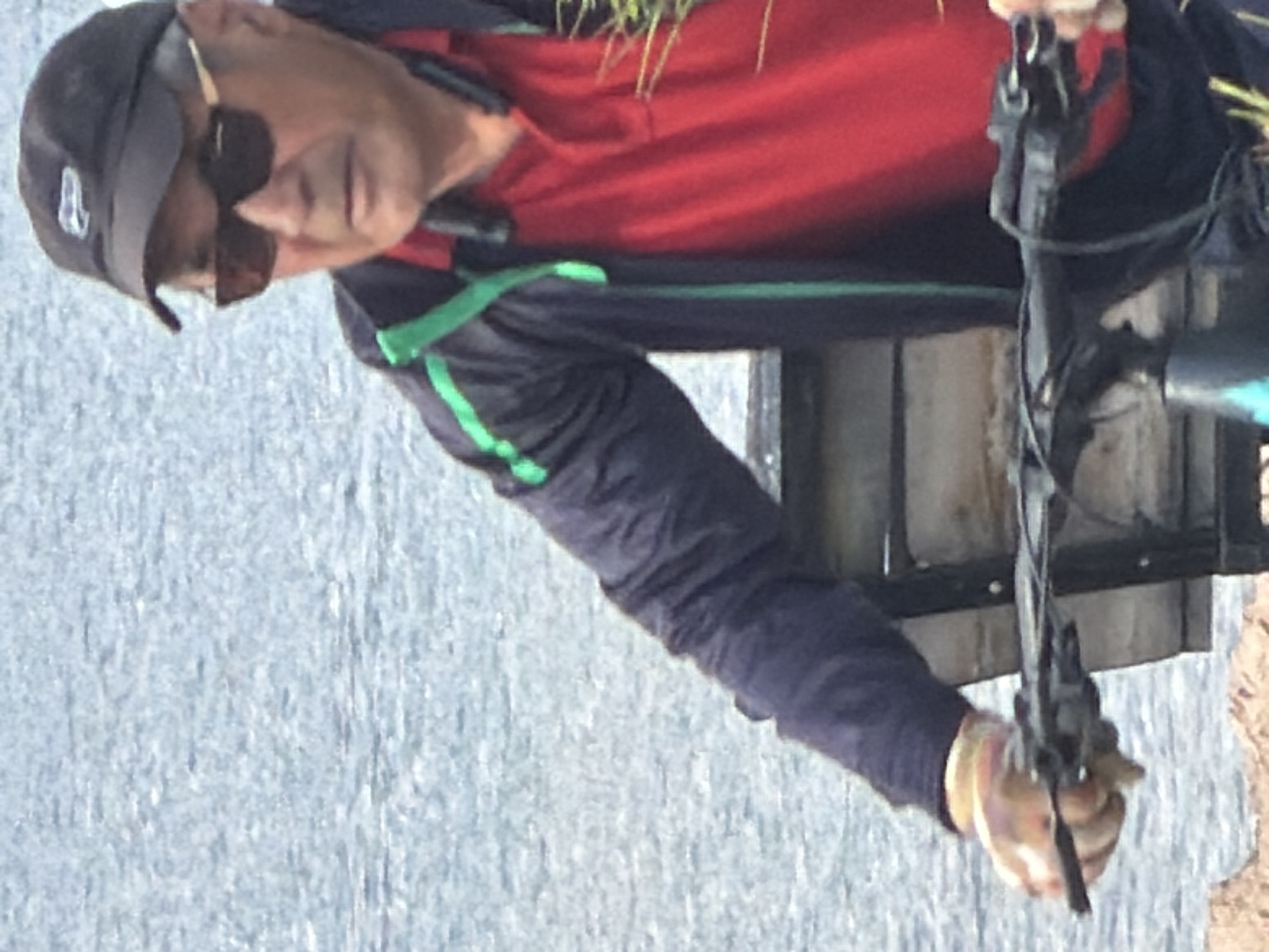 A man wearing a black baseball cap, sunglasses, a red polo shirt, and a dark blue jacket with green trim is sitting on a park bench. He is wearing gloves and holding onto the handlebars of a bicycle. The background is a body of water, possibly a lake or ocean, with a rocky shoreline visible in the foreground. The scene appears to be outdoors in daylight.