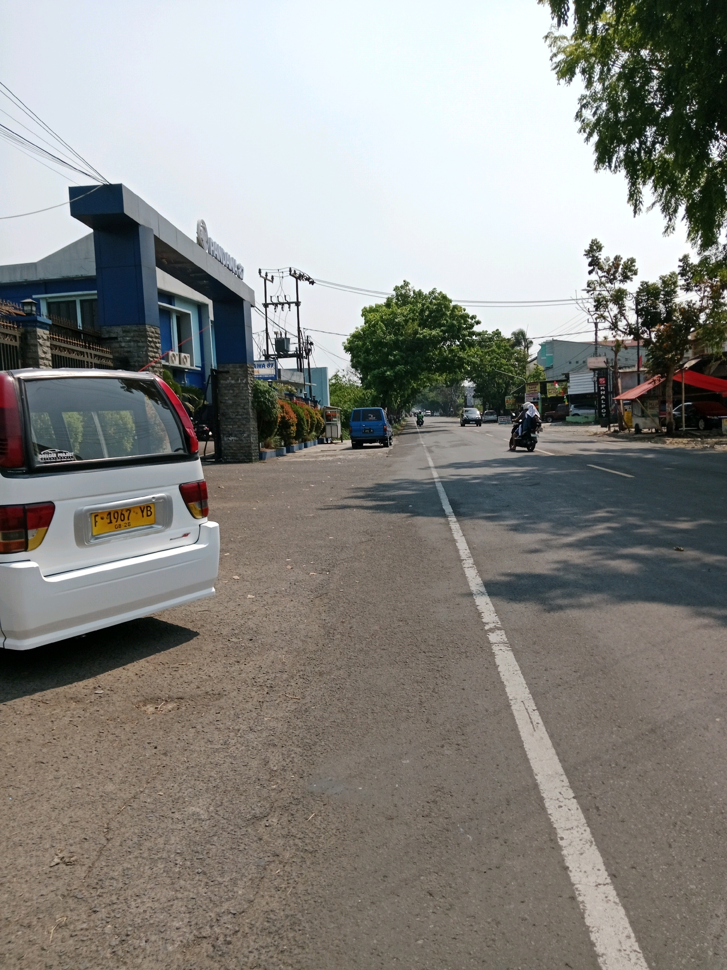 This is a medium-angle shot of a street scene in Muka, Indonesia. On the left, a white car is parked on the side of the road, facing away from the camera. The car has a yellow license plate that reads "F-1967 YB". Behind the car is a building with blue and gray accents, and an arched entrance with the text "PANDAN..." visible on it. Several electrical poles and wires are present in the background.
The road itself is asphalt and has a white dividing line down the center. Several vehicles are on the road, including a blue van further down on the left and a motorcycle with two riders in the middle of the road, heading away from the camera. Trees with green foliage line the sides of the road, and on the right, there are several small shops with red awnings and various stalls. The sky is bright and clear, indicating a sunny day.