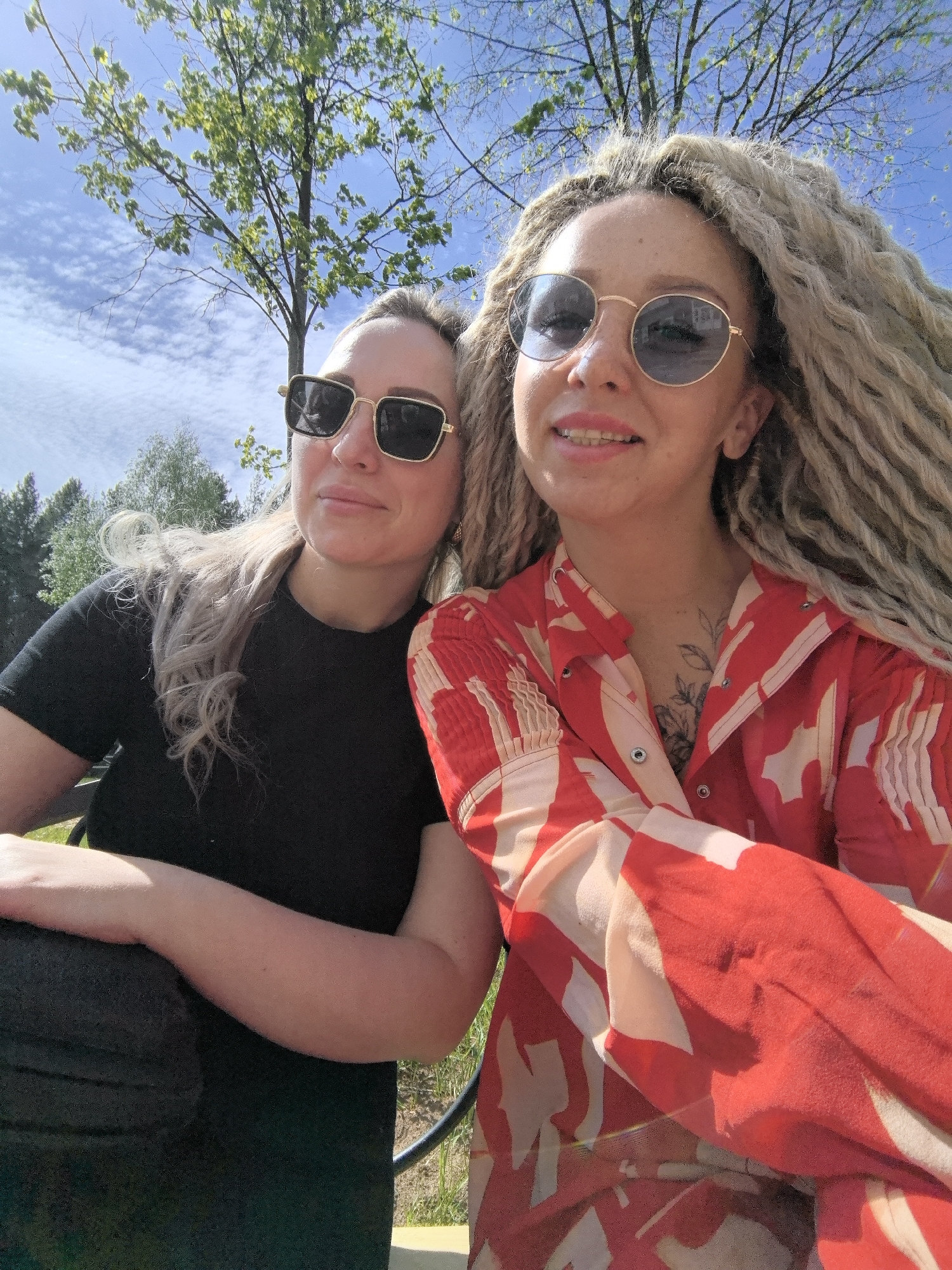 Two women pose outdoors. The woman on the left wears a black t-shirt and gold-rimmed square sunglasses. The woman on the right wears a red and white patterned button-down shirt and round, gold-rimmed sunglasses. She has long, blond dreadlocks and appears to have braces. A tattoo is visible on her chest. The background includes a blue sky with sparse clouds and green trees. The location may be near trees in the city of Chaikovsky, Russia.
