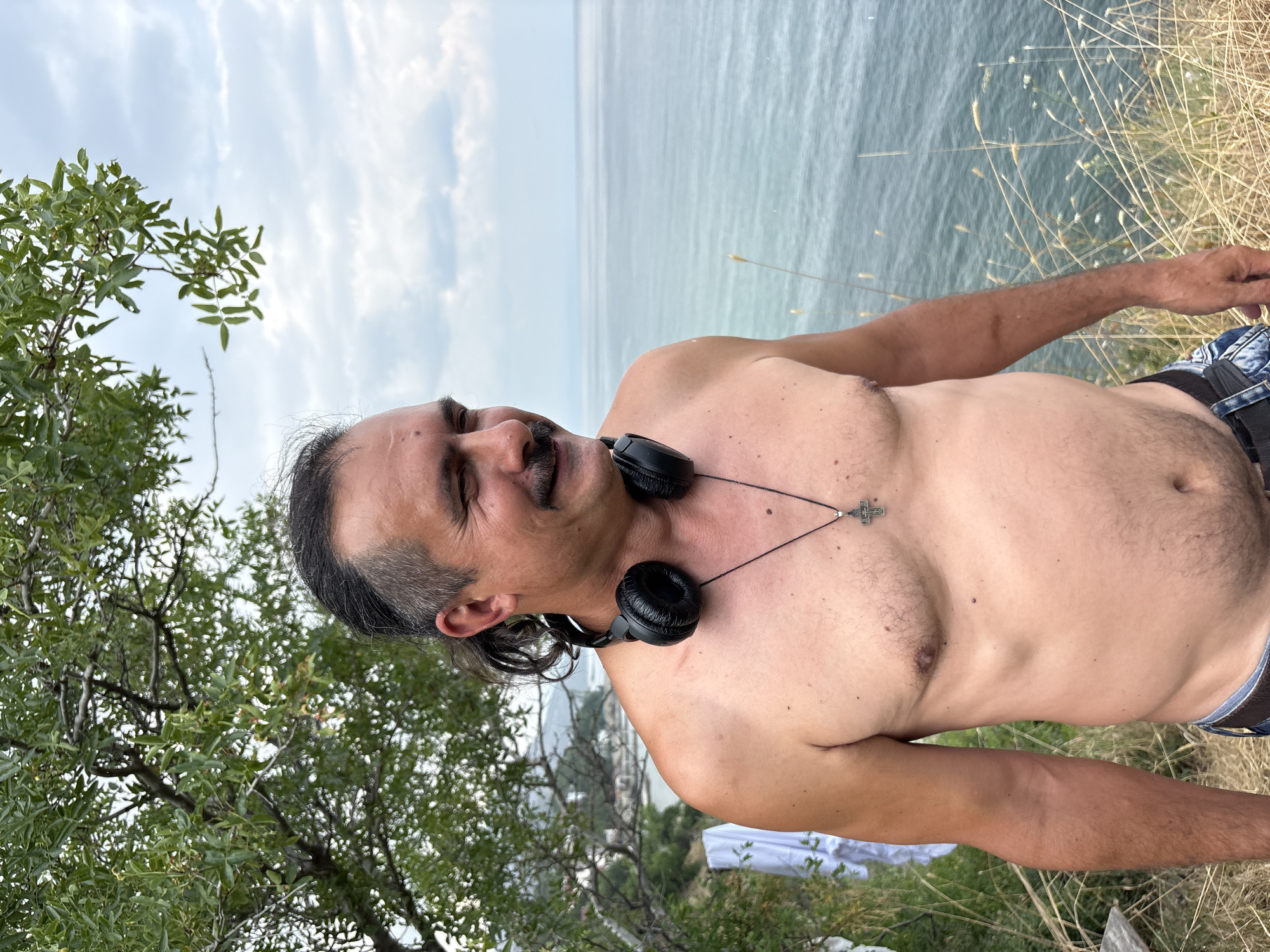 A shirtless man with a mustache, his hair tied back in a ponytail, stands on a grassy cliff overlooking the sea. He is wearing black headphones around his neck and a silver cross pendant. The background shows a body of water under a cloudy sky, with some trees and buildings visible in the distance along the coastline. The image is captured outdoors in what appears to be a natural setting.