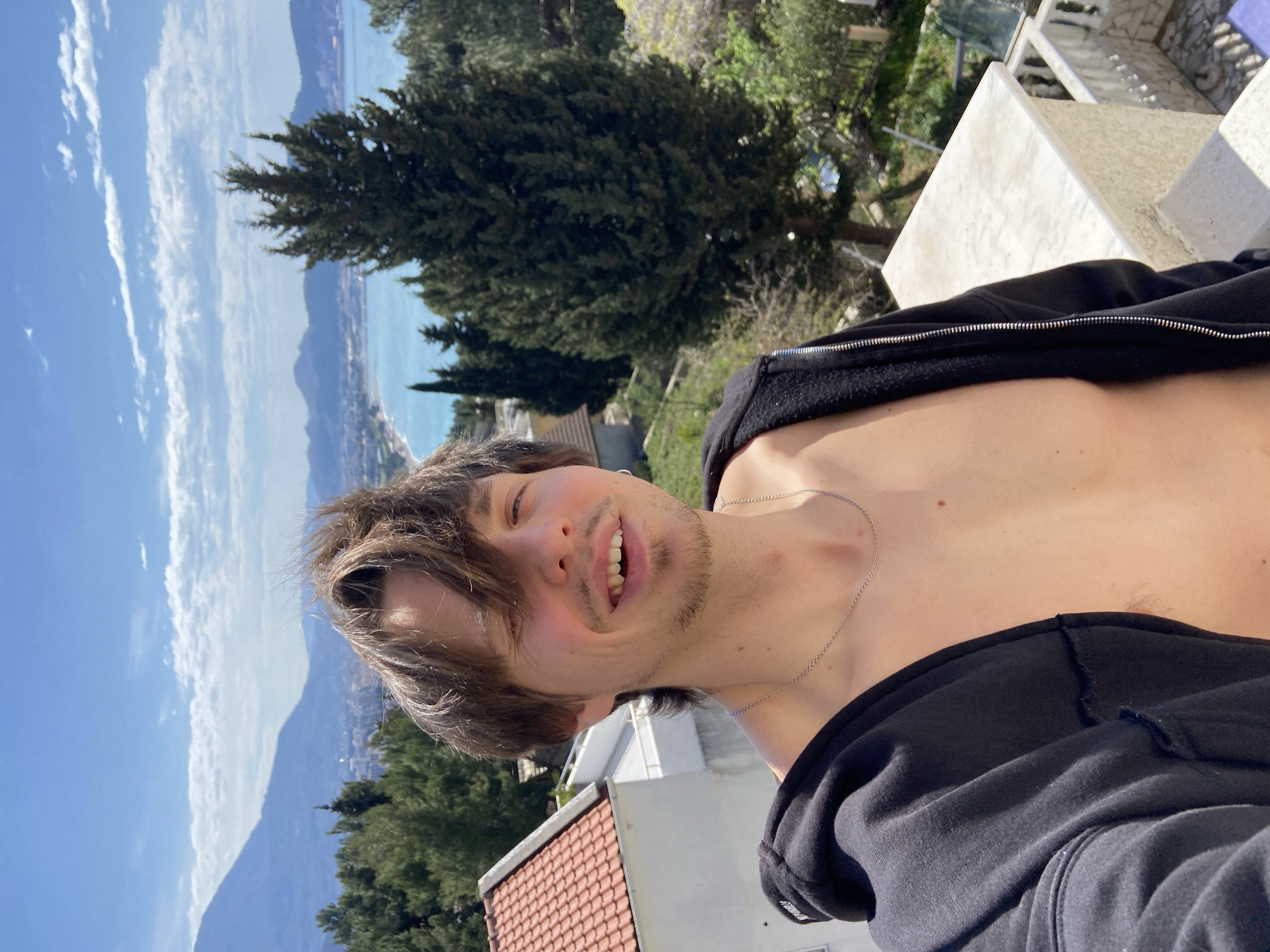 A male with brown hair and a slight beard stands on a rooftop, wearing a black hoodie open to reveal his bare chest and a silver chain necklace. The background shows a coastal view with the city of Šušanj, Montenegro, below a mountainous landscape under a blue sky with scattered clouds. A red-tiled roof and white structures are visible near the man. Trees are present in the mid-ground.