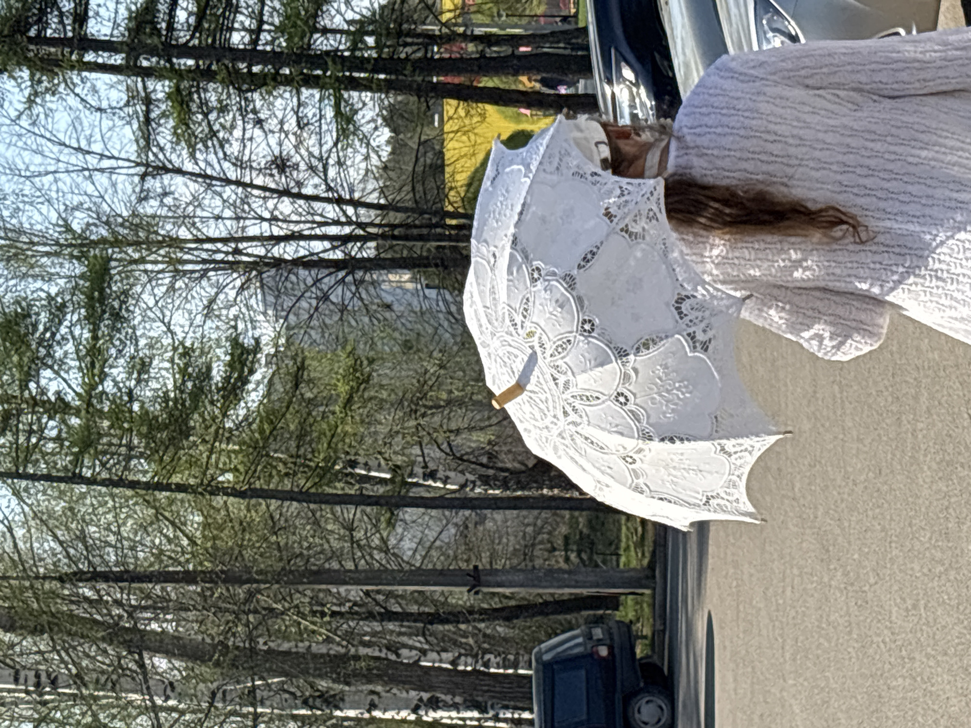 A woman, facing right and dressed in white, holds a white lace umbrella on a paved surface. A dark-colored vehicle is parked to the left of her. Beyond the vehicle, a metal fence runs horizontally across the scene. Behind the fence are trees with budding leaves, creating a dense background. The buildings in the background suggests the city of Chaikovsky, Russia.
