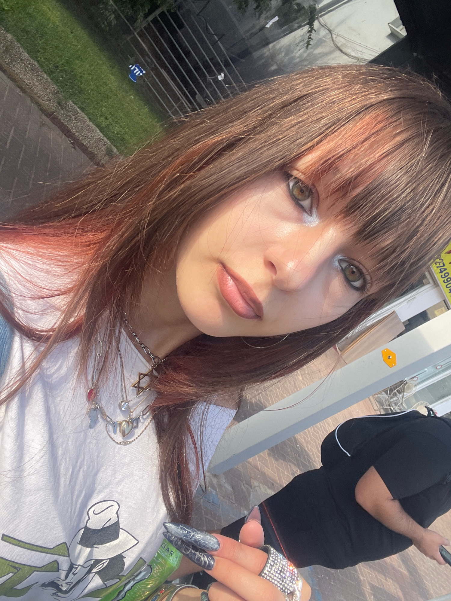 A young woman is taking a selfie outdoors in what appears to be an urban setting. She has long brown hair with pink highlights and is wearing a white t-shirt with a graphic print, layered silver necklaces, and several rings on her fingers, including a large rhinestone ring. Her nails are long and painted with a dark, textured design. In the background, a person in a black hoodie and pants is visible. The ground is paved with a mosaic-like pattern, and a metal fence with green grass behind it is to the left. A sign with Hebrew text is visible in the upper right corner of the image. The lighting suggests it is daytime. Given the Hebrew text on the sign, the location is likely in Israel, possibly Raanana, though specific landmarks are not identifiable from the image itself.