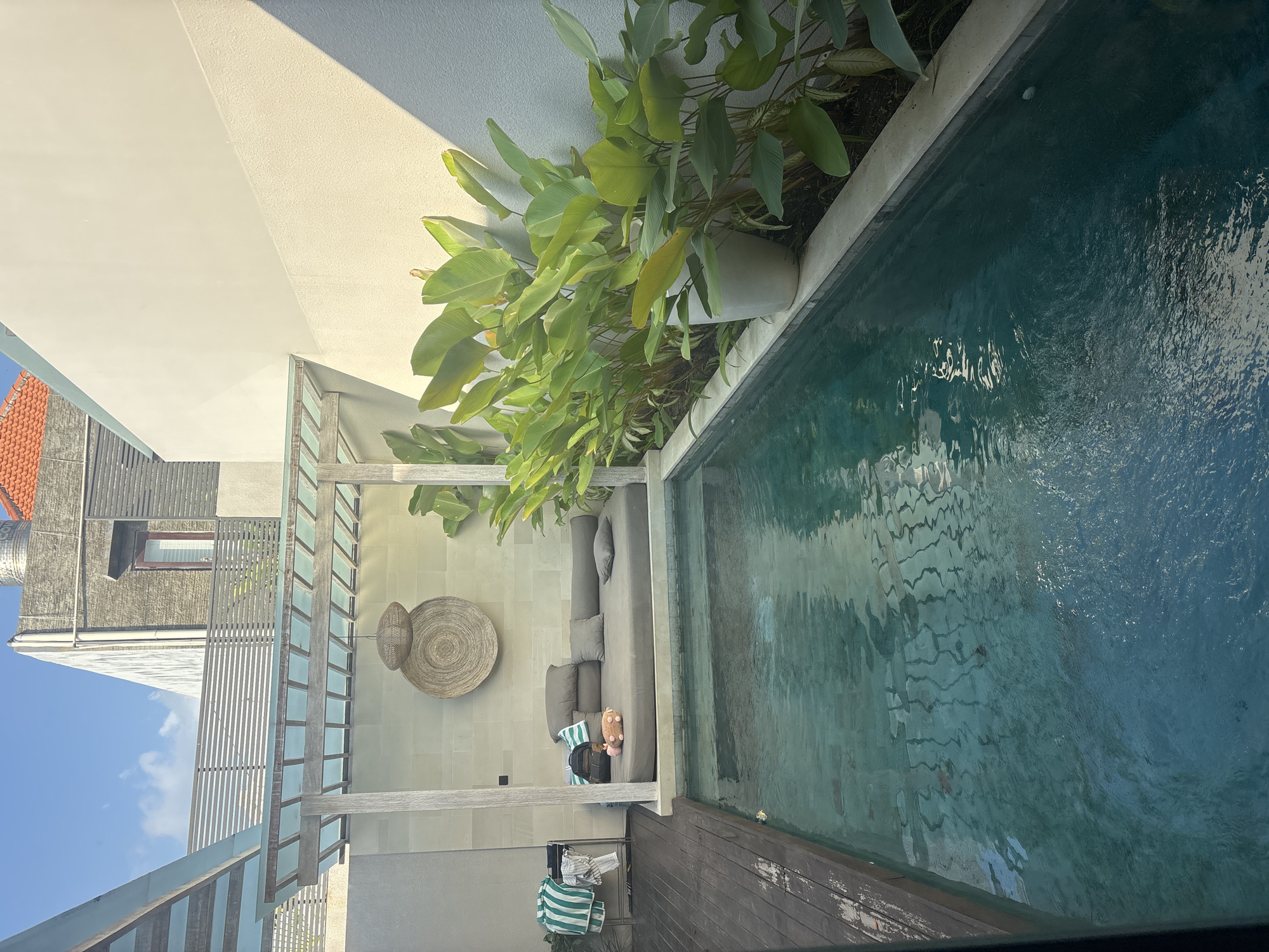 This is a photograph of a private swimming pool area within a building complex in North Kuta, Indonesia. The pool itself is dark blue, with visible tiling on the bottom. To the left of the pool is a wooden deck with a drying rack holding striped towels and clothing. Adjacent to the pool is a covered daybed area with a grey mattress and several cushions, featuring decorative woven wall hangings and a backpack. In the background, lush green plants line the edge of the pool, in front of a light-colored wall. The overall impression is one of a tranquil, outdoor living space, likely a resort or private villa.