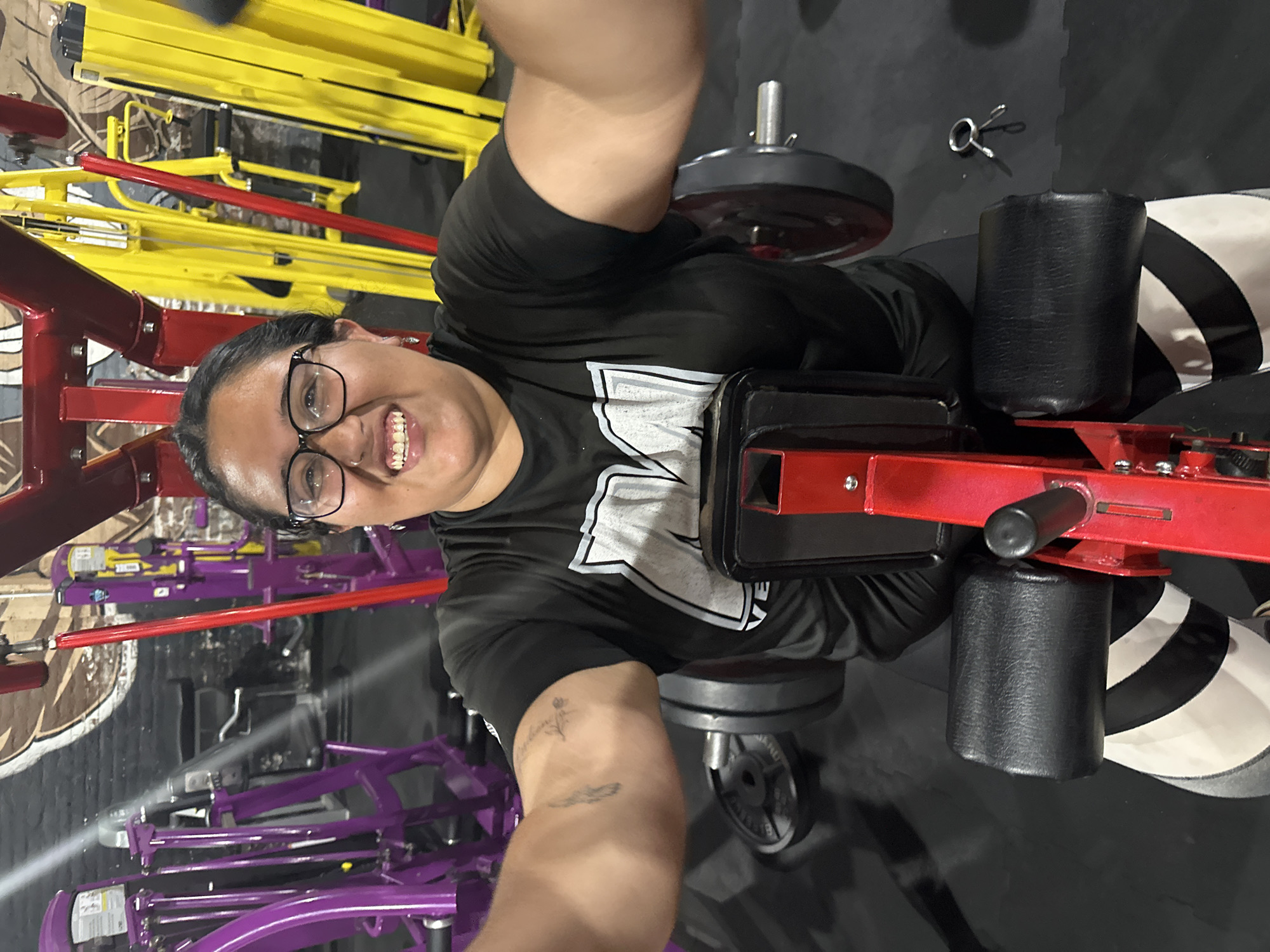 A smiling person with glasses is in a gym, likely in Arica, Chile, although there is no visual confirmation in the image to confirm the location. They are using a leg extension machine with weights loaded on it. The machine has red and black components, and there are other gym machines visible in the background, including purple and yellow equipment. The gym has a black floor. The person is wearing a black t-shirt with a white logo. A tattoo is visible on the person's arm.