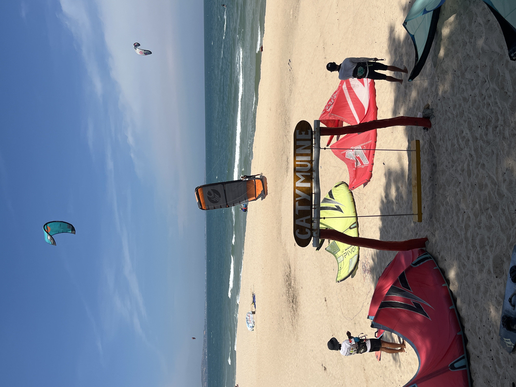 The image captures a sunny day at a beach in Phu Thuy Ward, Vietnam, likely a location frequented by kitesurfers. In the foreground, several kitesurfing kites are laid out on the sand, with two individuals present. One person, wearing a black hat and a white t-shirt with graphics, stands near a large red and black kite. Another person, wearing a black baseball cap and a white long-sleeved shirt, stands near a red and white kite. A wooden swing set with a yellow seat is positioned between the kites. Above the swing set hangs a wooden sign with the words "CATY MUINE" carved into it. In the middle ground, more kitesurfing kites are visible on the sand, and a kitesurfer is in the water. The ocean stretches to the horizon under a bright blue sky with scattered white clouds. Further in the distance, several kites are flying, and a few people are on the beach. The overall scene suggests a vibrant and active kitesurfing spot.