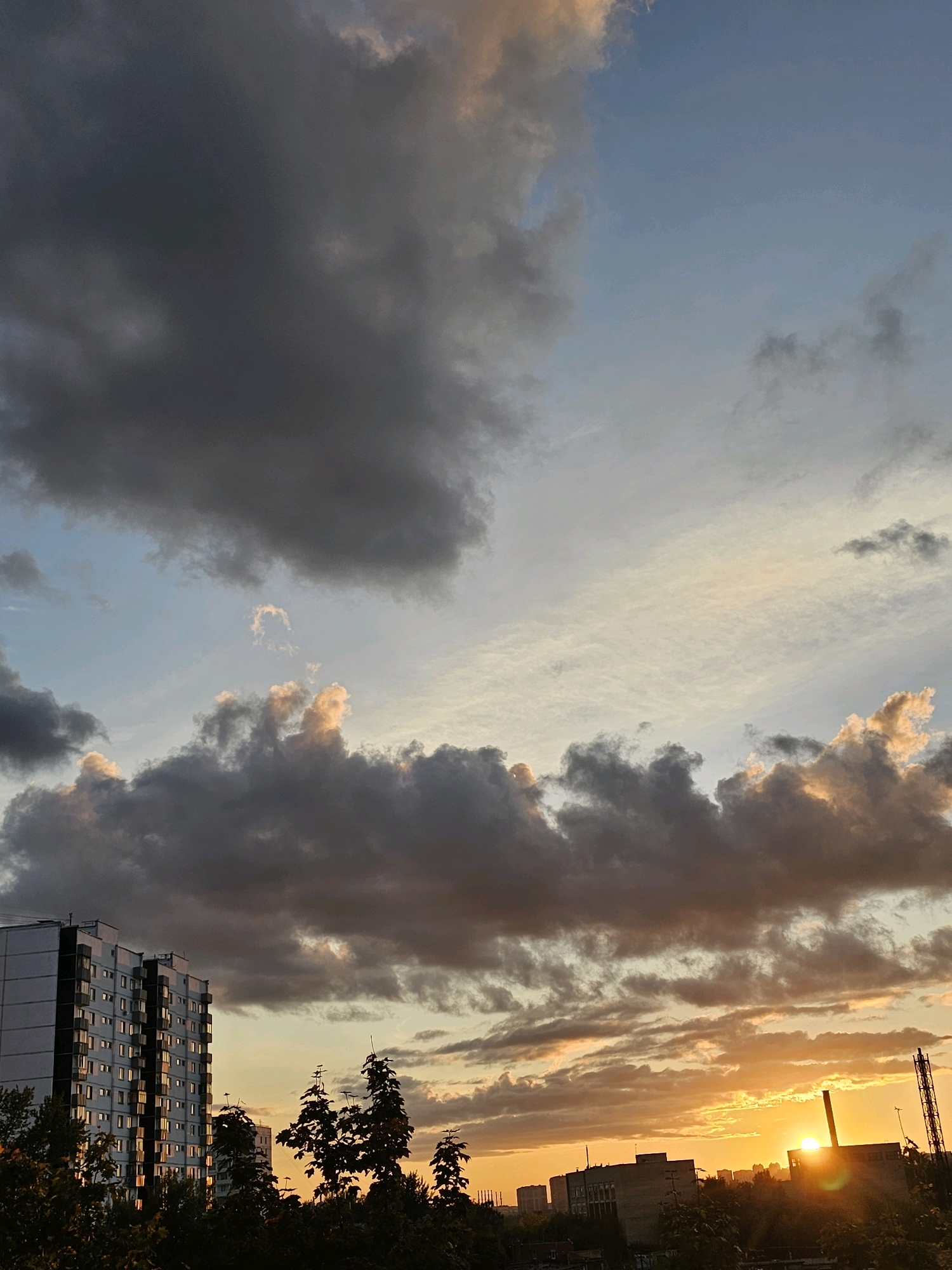 The image captures a sunset over the city of Khimki, Russia. Dark, heavy clouds dominate the upper left portion of the sky, transitioning to lighter, wispy clouds with hints of orange and pink closer to the horizon. The sun is a bright orb peeking through the clouds on the right side of the frame, casting a warm glow over the scene.
In the foreground on the left, a tall, white apartment building stands prominently. Its windows reflect the warm light of the sunset. Silhouetted trees and greenery fill the lower portion of the image, obscuring the bases of the buildings and creating a natural foreground. In the distance, on the right, other buildings and industrial structures are visible, silhouetted against the vibrant sunset. A tall, lattice-like structure, possibly a radio tower or industrial chimney, is also present on the right side. The overall atmosphere is one of a dramatic and colorful end to the day.