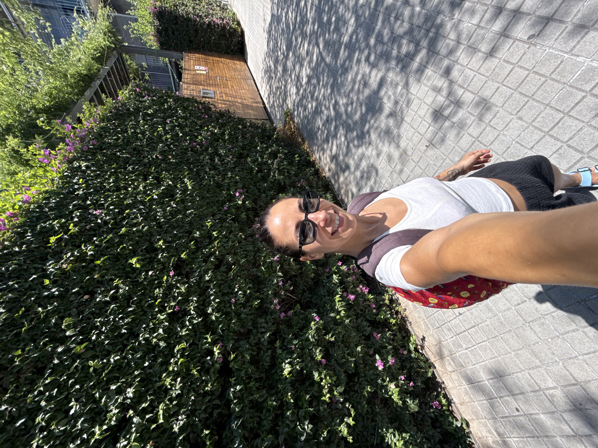 A woman with dark hair, wearing sunglasses, a white tank top, and black shorts, takes a selfie while walking on a tiled sidewalk in Barcelona, Spain. She is carrying a red backpack. The sidewalk is shaded by trees, creating a dappled light effect. To her left is a dense green hedge with small pink flowers, and to her right is a wooden fence and a building. The scene appears to be in an urban area with a modern architectural style.