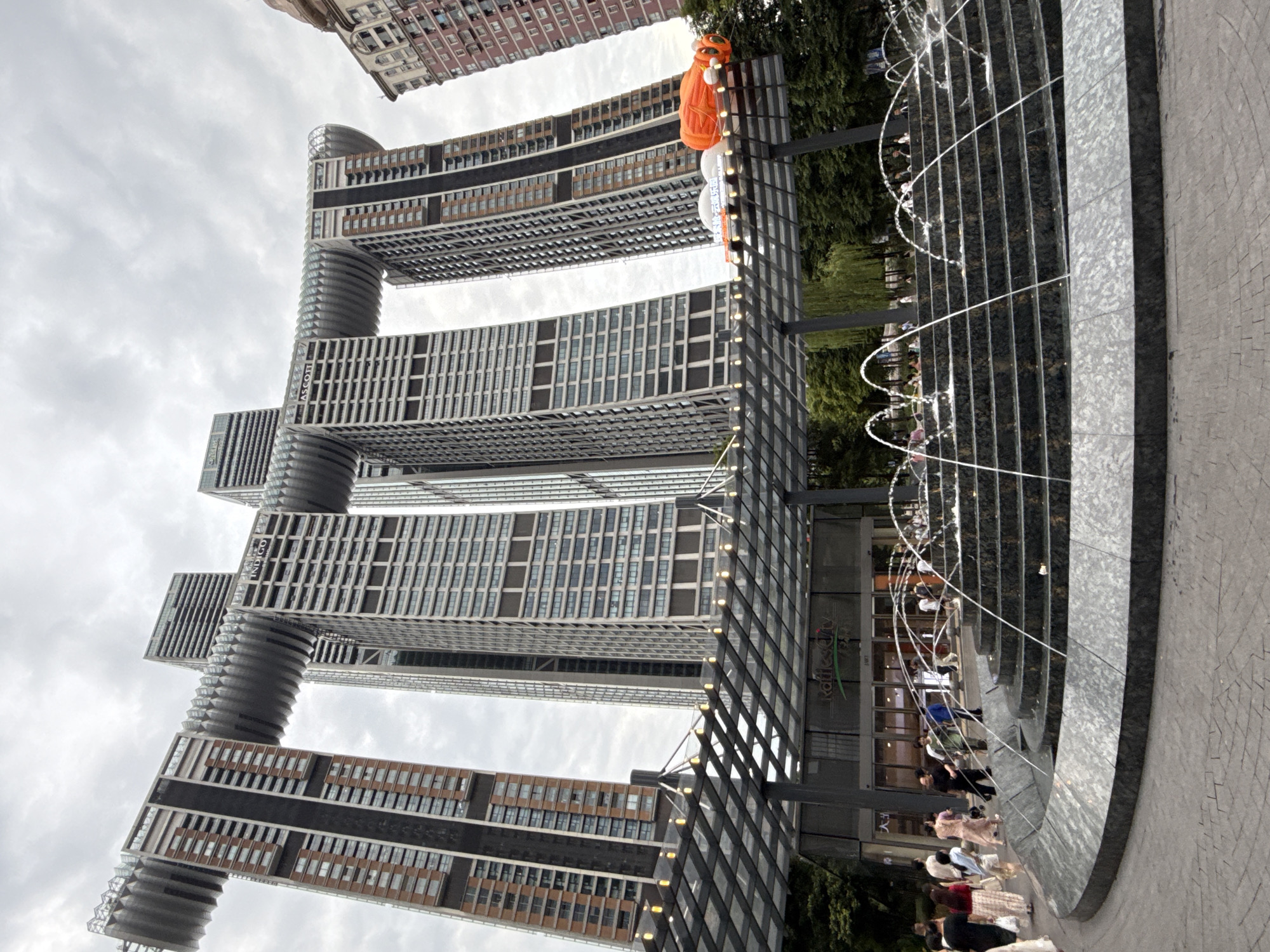 The image displays the Raffles City complex in Chongqing, Yuzhong District, China. The complex features four interconnected skyscrapers with a distinctive horizontal element connecting them at the top, resembling a ship or a crystal. In the foreground, a multi-tiered fountain with cascading water is visible, surrounded by people walking on a paved plaza. The buildings are clad in a combination of glass and brick facades, and the sky is overcast. An orange, inflatable object, possibly a decoration, is positioned on a walkway above the fountain, near the buildings. The overall scene captures a busy urban environment with modern architecture and public amenities.
