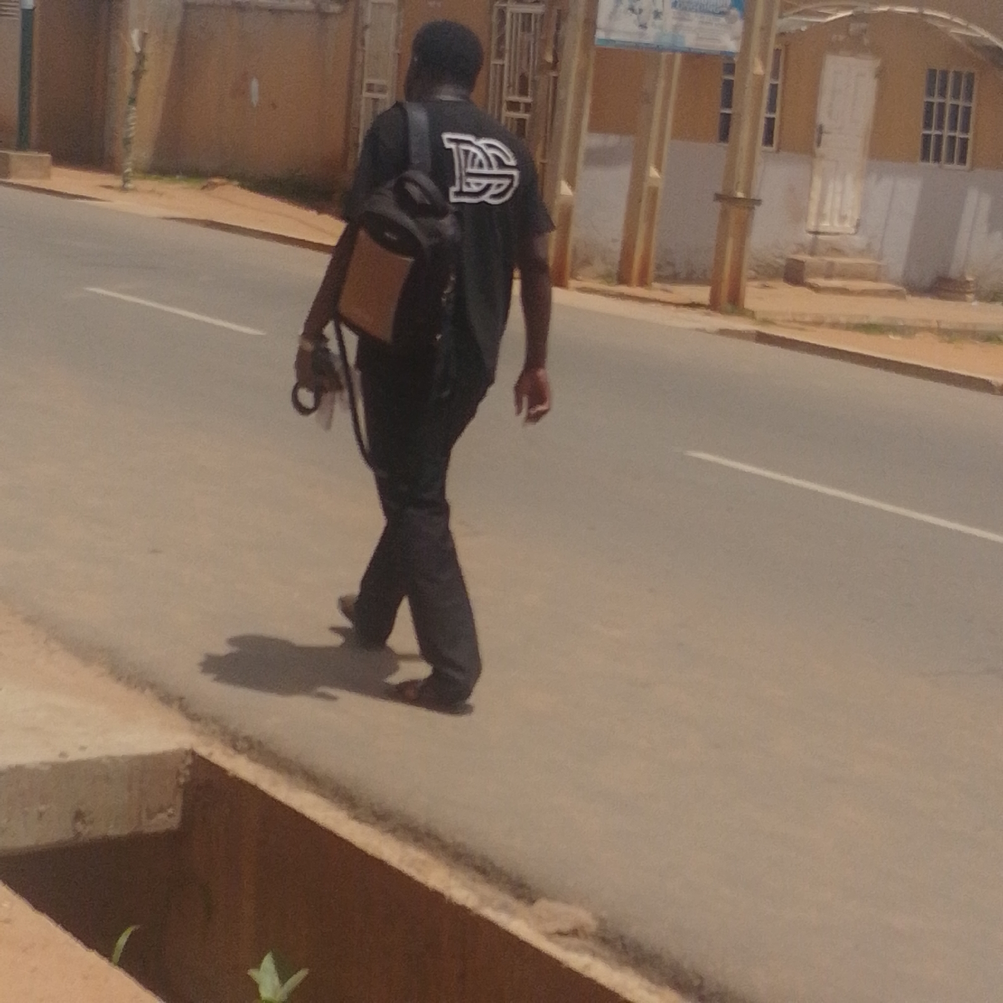 A man is walking away from the camera on a paved road in Tempure, Nigeria. He is wearing a black t-shirt with a white logo on the back, dark pants, and dark shoes. He has a backpack on his left shoulder and is carrying a camera in his right hand. The road has a white line marking the center. To the left of the road, there is a concrete structure with some plants growing in it. In the background, there is a building with a yellow facade and a white door. There are windows on the building, some of which have bars.