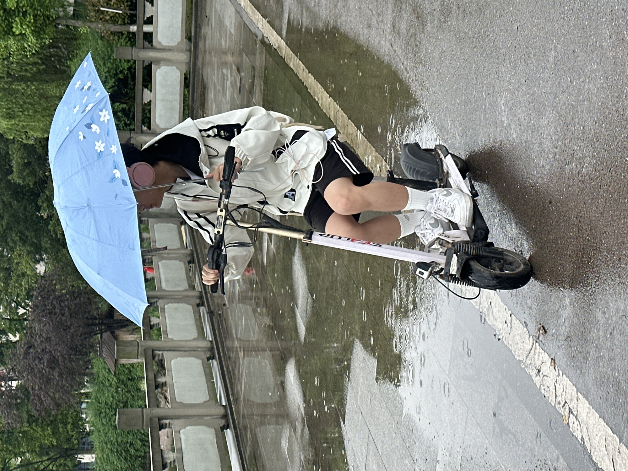 In Jiangjin, China, a person rides a white electric scooter on a wet, light grey paved surface, with rain actively falling. The rider holds a light blue umbrella with a pattern of white flowers. The individual is wearing a white jacket with black stripes, black shorts, white socks, and white sneakers. Pink headphones are visible on their head. A low stone wall with decorative rectangular cutouts runs parallel to the scooter. Green vegetation is visible behind the wall.
