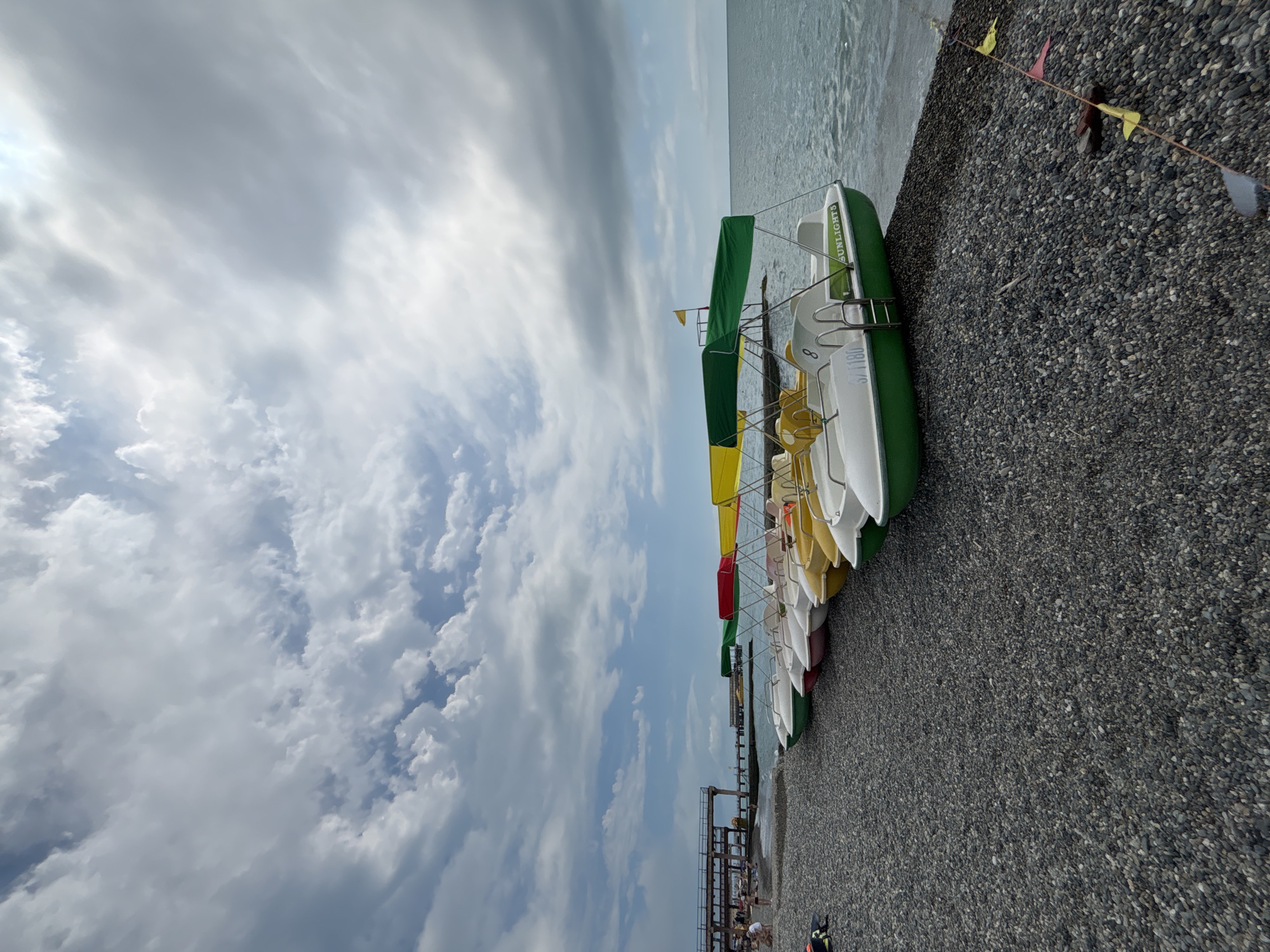 The image depicts a beach in Sochi, Russia, on a cloudy day. A row of paddle boats, in various colors including white, yellow, red, and green, are lined up on the pebble shore, with their bows facing the sea. The water of the Black Sea is calm and appears slightly choppy. In the background, a pier extends into the water, with a few people visible on it and on the beach. The sky is filled with a mix of white and gray clouds, with some patches of blue visible. A string of small, colorful flags is partially visible in the foreground, running along the edge of the beach. One of the paddle boats, a green one in the foreground, has "SUNLIGHT 5" written on its side. The overall scene is one of a quiet, overcast day at the beach.