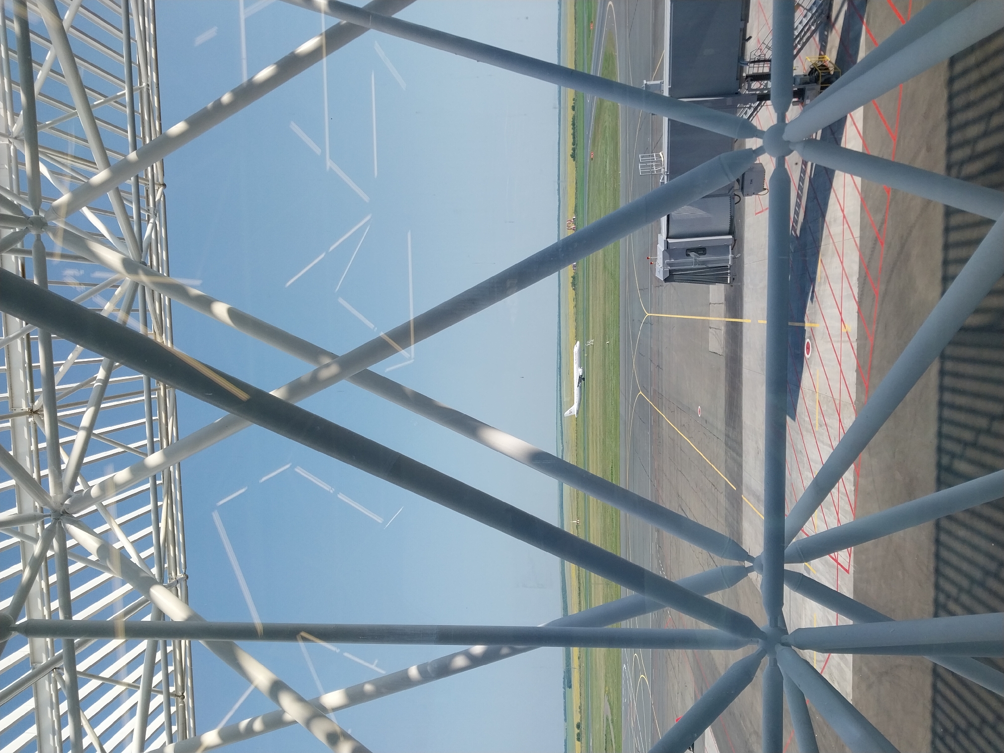 An exterior shot captures a clear blue sky above an airfield. In the foreground, a geometric lattice structure made of white metal beams obstructs the view. The lattice is composed of intersecting diagonal and horizontal beams that converge at nodal points. Beyond the structure, an airplane is visible on the tarmac, positioned on the left side of the frame. The airfield features a paved surface with painted lines and markings, and to the right, a jet bridge extends towards the airplane. The background shows a flat landscape with green fields and some scattered trees under a bright sky. The image appears to be taken from inside an airport terminal or a connecting walkway. Based on the context of the Orenburg airport, this scene likely depicts the area around Orenburg Tsentralny Airport.