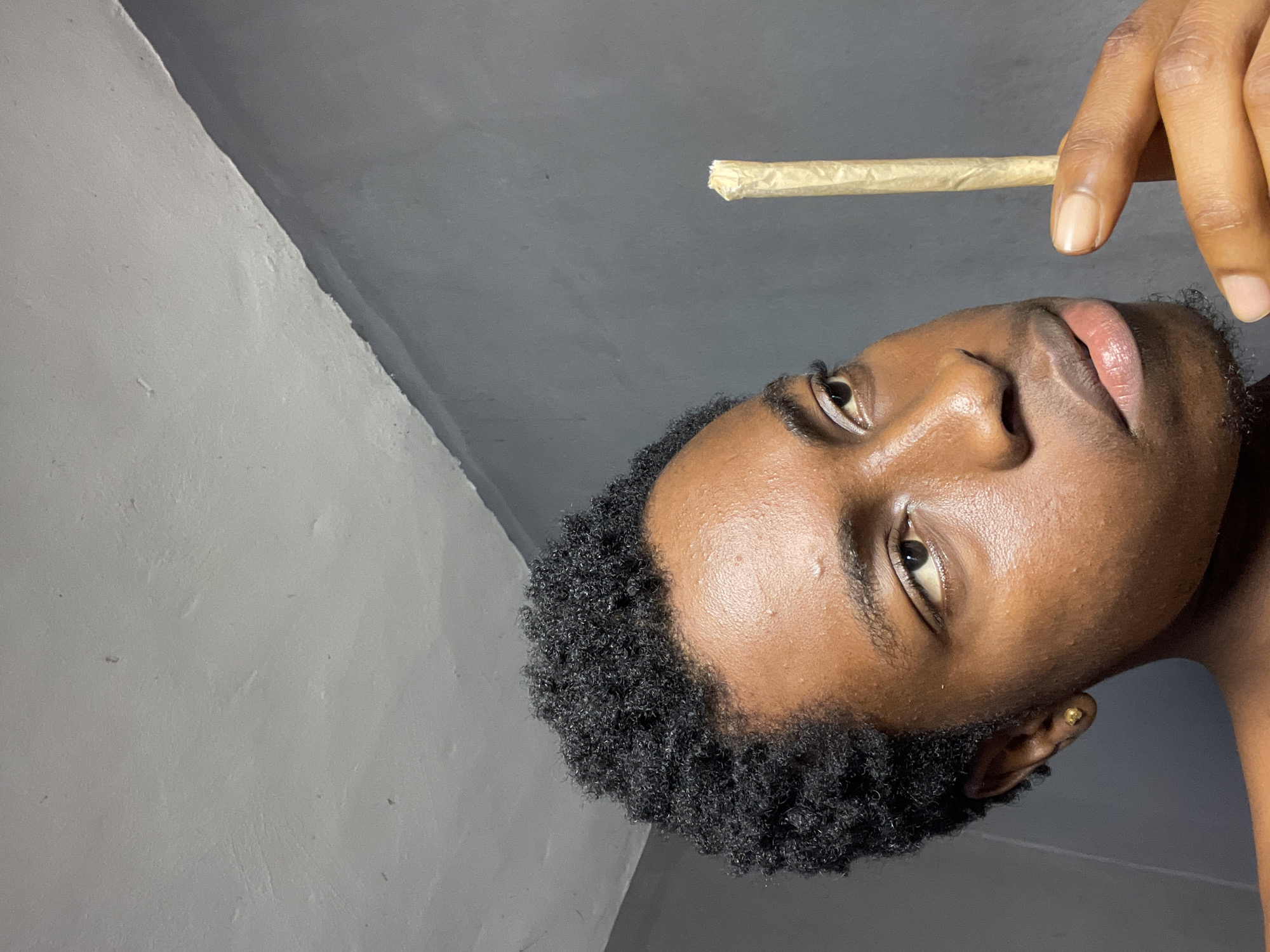 A close-up, angled shot depicts a Black male holding a rolled cigarette or joint. The man has short, dark, tightly curled hair. His skin is a medium brown with visible pores. He has a gold earring in his left ear. His lips are full and appear slightly glossy. He is holding the rolled cigarette between his fingers. The background consists of a gray wall in a room. The location is unspecified in the city of Nkpor, Nigeria.