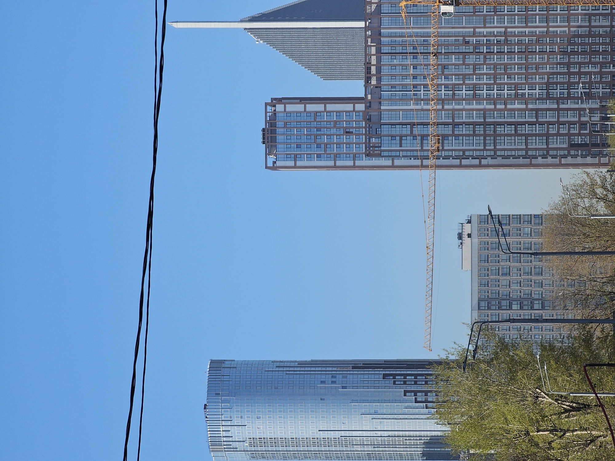The image shows a view of buildings and construction in Moscow, Russia. In the foreground are several power lines against a clear blue sky. At the bottom is a modern, curved glass building reflecting the sky. Above it, behind some bare trees and streetlights, is a more traditional residential building. Further up, there is a taller modern building with a construction crane beside it. At the top of the frame is a section of another building with a flat, projecting roof.