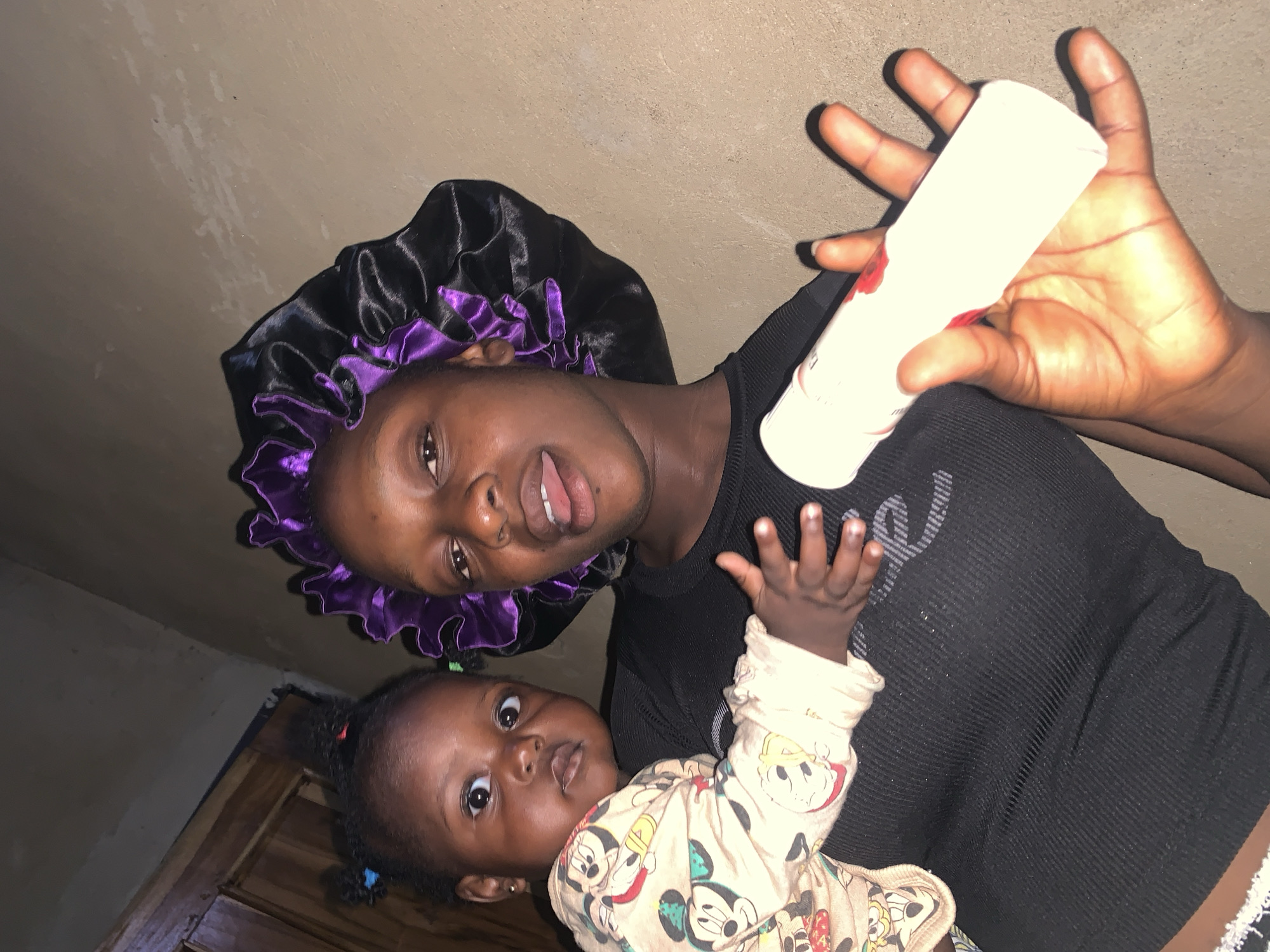 In Agban, Nigeria, a young Black woman wearing a black ribbed tank top and a purple and black satin bonnet holds a baby. The baby, wearing a long-sleeved shirt with a Mickey Mouse print, is cradled in the woman's arms. The woman is sticking her tongue out, and her right hand is extended forward, holding a white cylindrical object with a red label. The background consists of a plain, textured wall. The lighting is dim, suggesting an indoor setting at night.