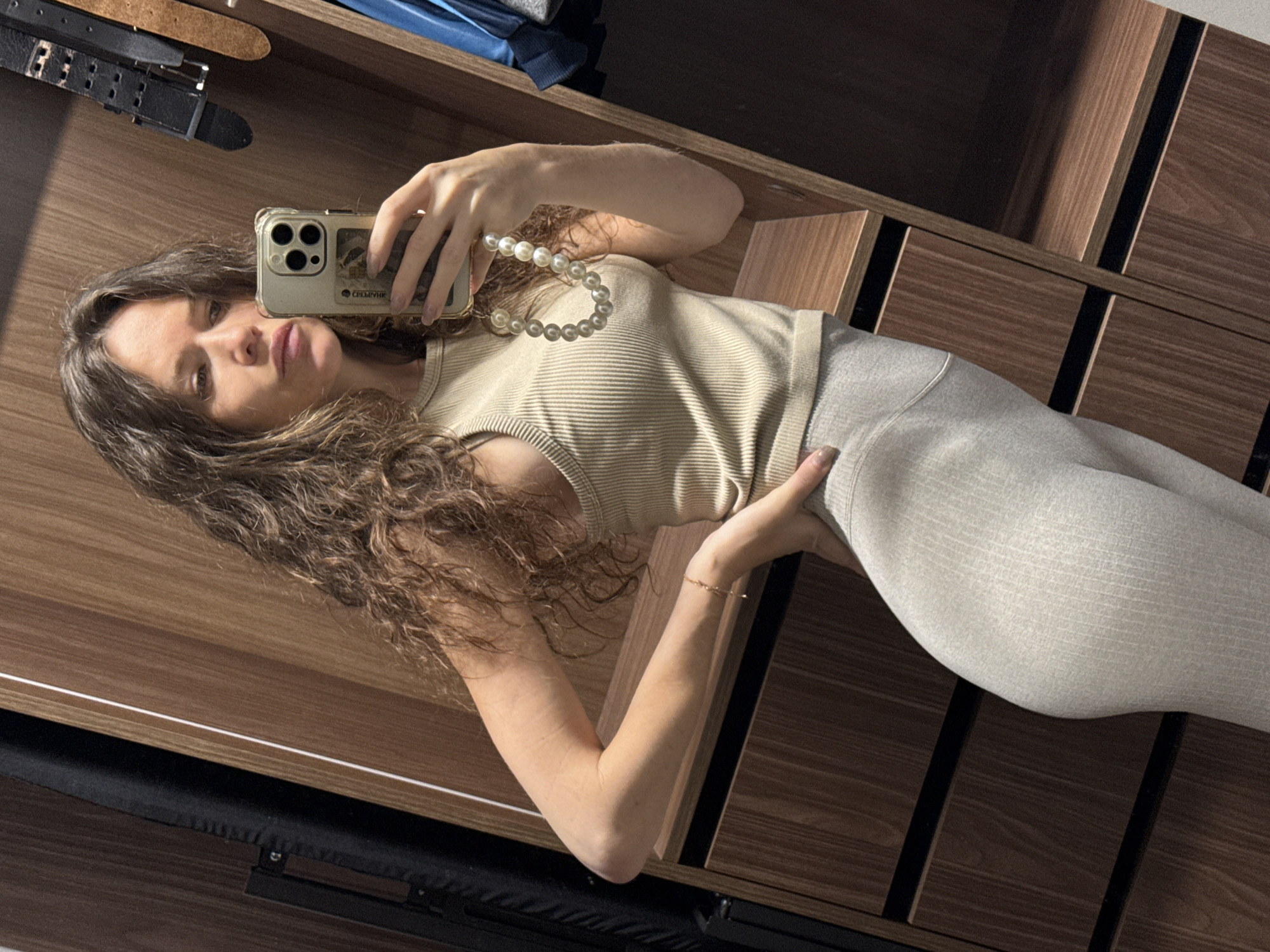 A woman takes a mirror selfie in an indoor setting, likely a dressing area or bedroom in Moscow, Russia. She has long, wavy brown hair and is wearing a light-colored, possibly beige or light grey, ribbed tank top and matching leggings. She holds a smartphone with a gold/clear case, which features three camera lenses. A pearl necklace is draped around her phone, and she wears a delicate bracelet on her left wrist. Her nails are manicured.
The woman looks directly at her reflection with a somewhat serious or contemplative expression, her lips slightly parted. The background, reflected in the mirror, displays wooden furniture, likely a wardrobe or a set of drawers, with dark vertical and horizontal accents. Above the furniture, several belts are visible, including a black one with a buckle and a brown one. A blue object is also partially visible in the upper left corner of the original image (reflected top right).
Natural light illuminates the scene, casting subtle shadows. On the back of her phone case, visible Cyrillic text clearly reads "СБЕРБАНК".