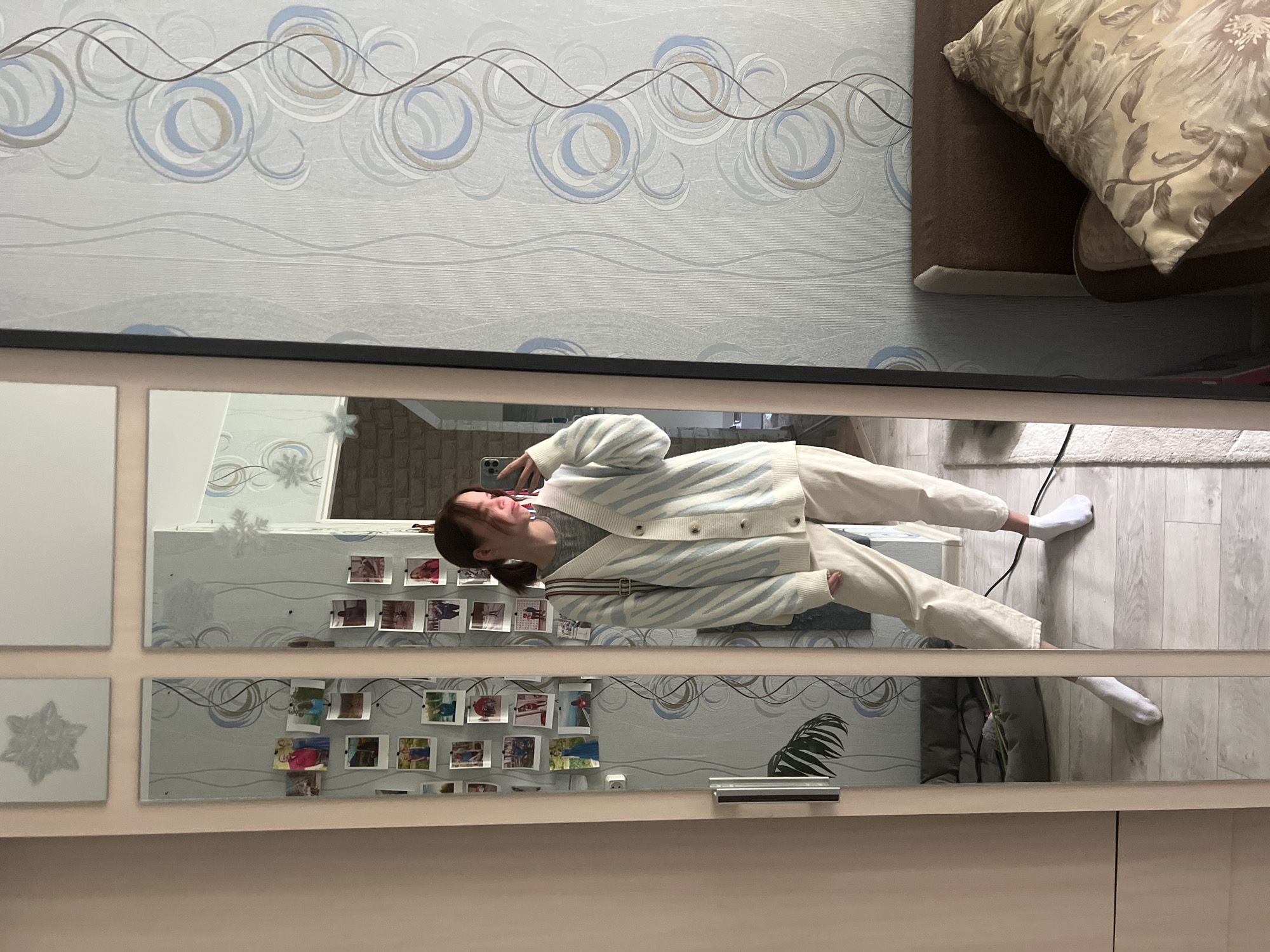 A person is reflected in a mirror, wearing a light blue and white striped cardigan, a grey shirt, and light-colored pants. They are holding a phone and taking a picture of themselves. The reflection is segmented by the design of the mirror, which consists of several square or rectangular panels. Some of the panels reflect a wall adorned with photographs and decorative elements like a leafy plant. The environment behind the person appears to be a room with light-colored walls and flooring. Based on the presence of specific wall patterns and furniture arrangements, this location may be a domestic interior in Chaikovsky, Russia. The overall scene depicts someone documenting their appearance in an indoor environment.