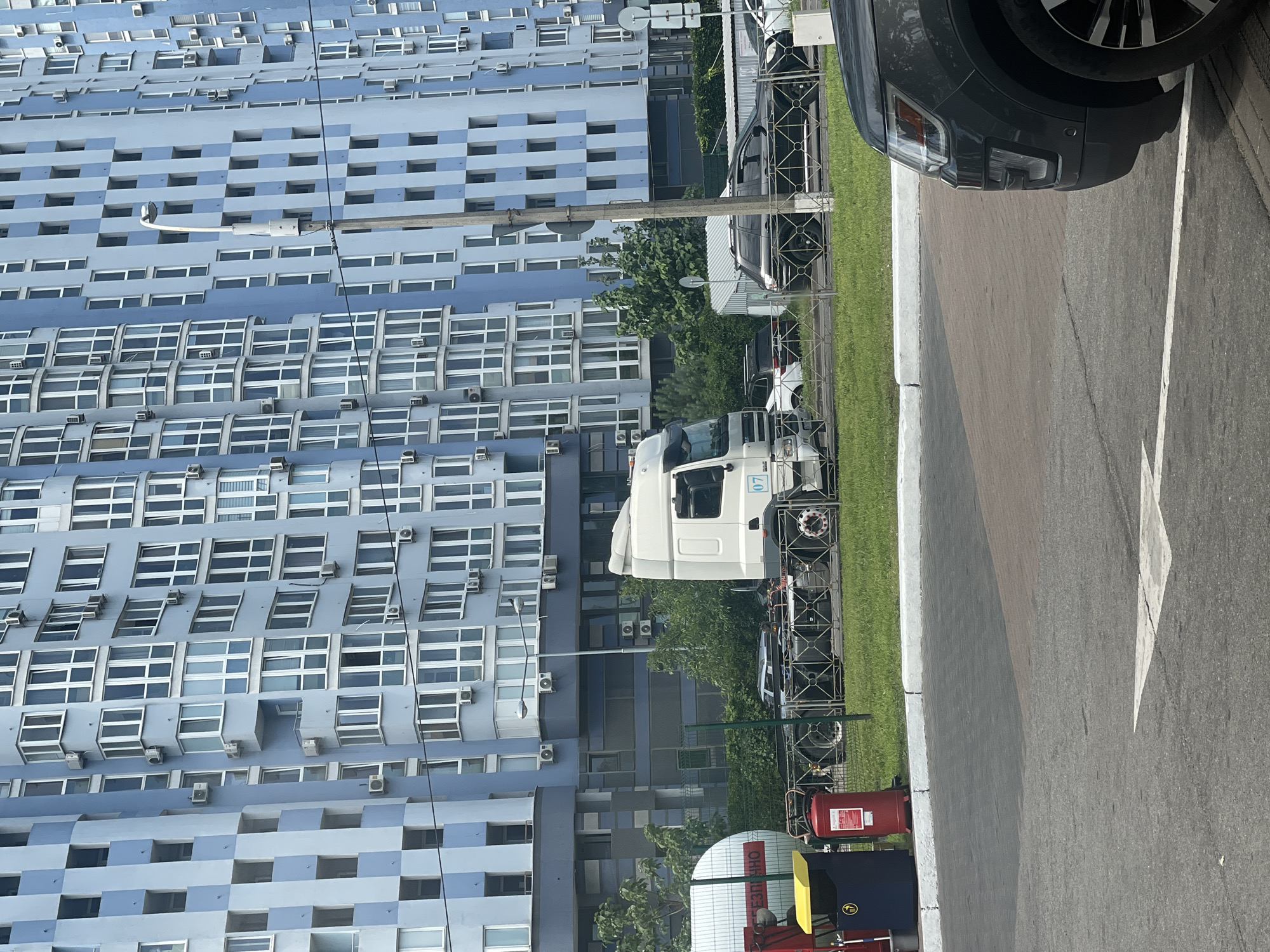 A white semi-truck is parked on the right side of a paved road in front of a blue and white checkered residential building in Kyiv, Ukraine. The building has numerous windows and balconies. To the left of the semi-truck, a large white cylindrical tank is visible, with a red fire extinguisher and a blue and yellow trash bin next to it. A metal fence separates the road from the grassy area in front of the building. Several other cars are parked or driving on the road, including a black SUV in the foreground. The sky is overcast.