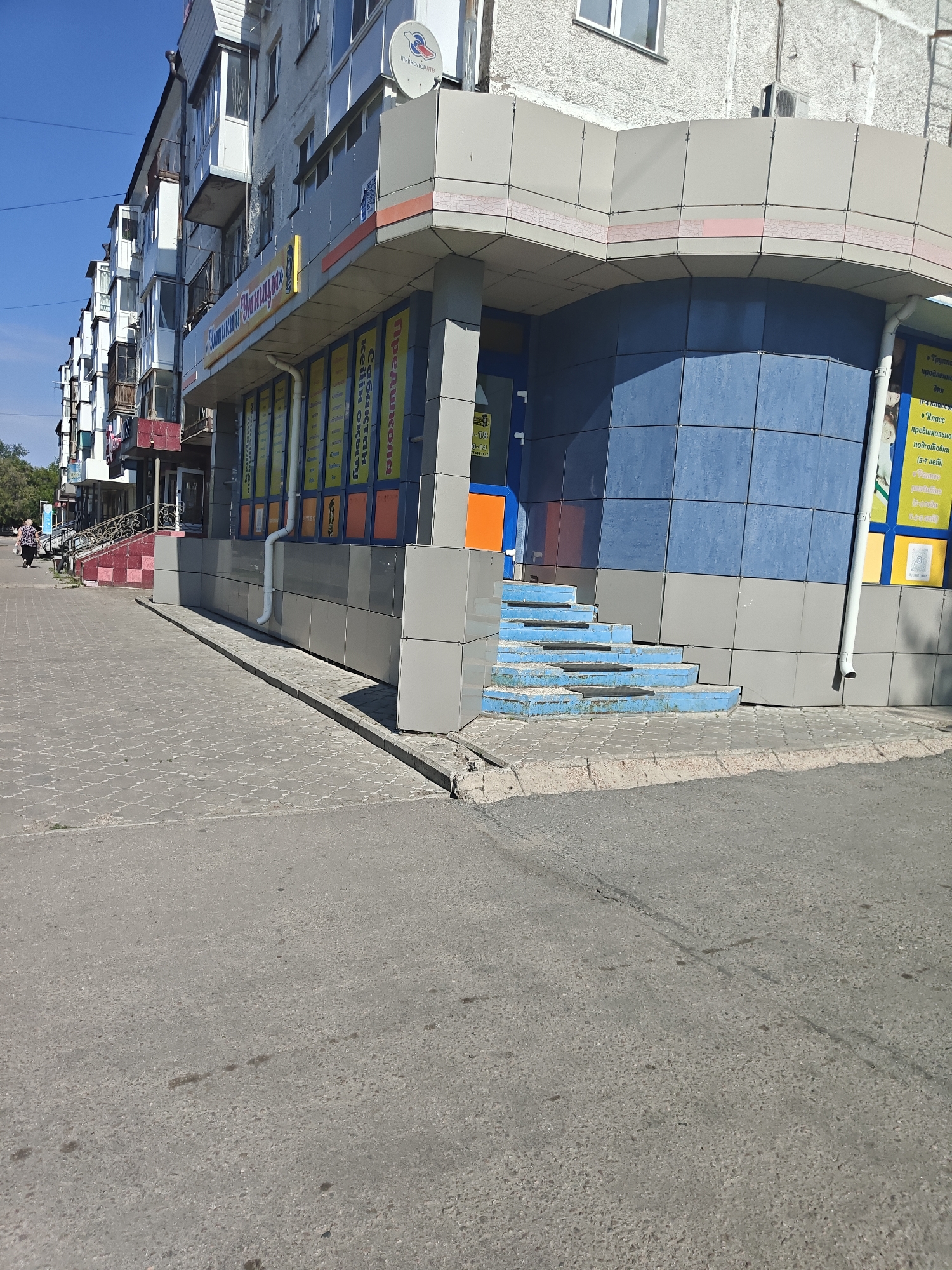 This is a daytime, eye-level, full shot of a building facade in Petropavl, Kazakhstan. The building appears to be a commercial establishment with a blue tiled curved section and an overhang. There are several signs visible. One sign in Cyrillic reads "Детский и Юношеский Центр" which translates to "Children's and Youth Center". There are also signs advertising classes for different age groups, for example, "1-4 классы" (1-4 grades) and "дошкольники (5-7 лет)" (preschoolers (5-7 years old)). A woman is walking on the sidewalk in the foreground. The sidewalk is paved with grey interlocking pavers, and there is a gray asphalt road in front of it. Blue steps lead up to the entrance of the building. The sky is clear and blue.