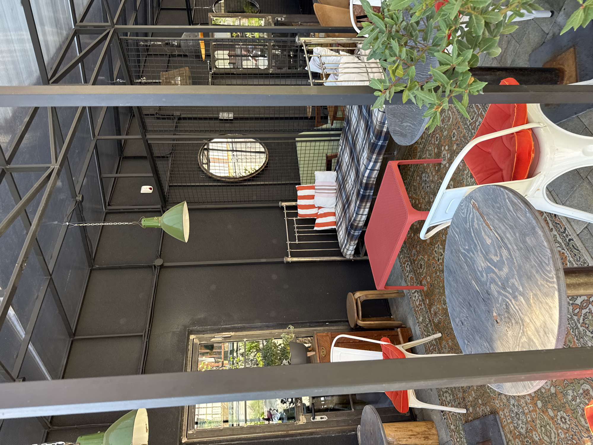 This image shows an indoor/outdoor space that appears to be a café or restaurant. The décor is eclectic, with a mix of industrial and vintage elements.
The space is enclosed by a metal frame structure, possibly a pergola or a covered patio. The walls are painted black. There are windows and an oval-shaped porthole-style window, allowing light into the space.
The furniture includes a bed (possibly for lounging), tables, and chairs with red cushions. The tables have wooden tops and the chairs are white. There is also a red table. A patterned rug covers the floor.
Hanging lamps with green shades illuminate the area. A potted plant is placed on a table.
The reflections in the windows show hints of the outside environment, but it is not possible to determine the precise location within Krasnaya Polyana from the image. There is no visible presence of people.